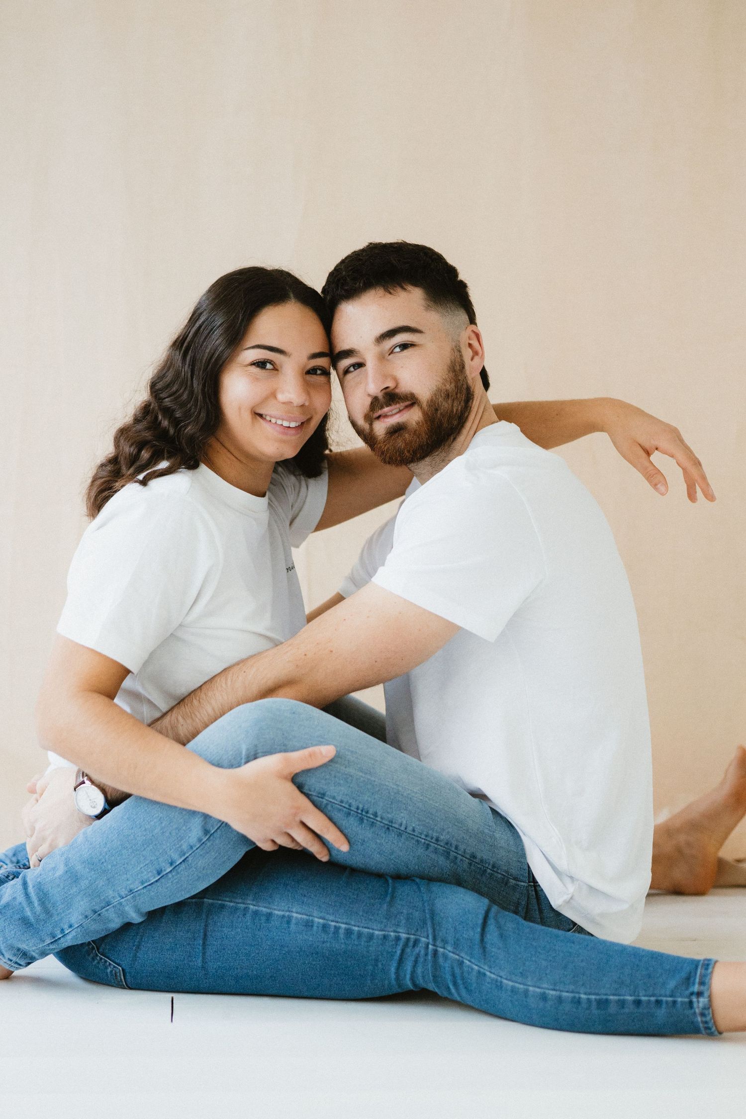 Couple in casual attire lying together on floor in romantic poses against neutral backdrop.