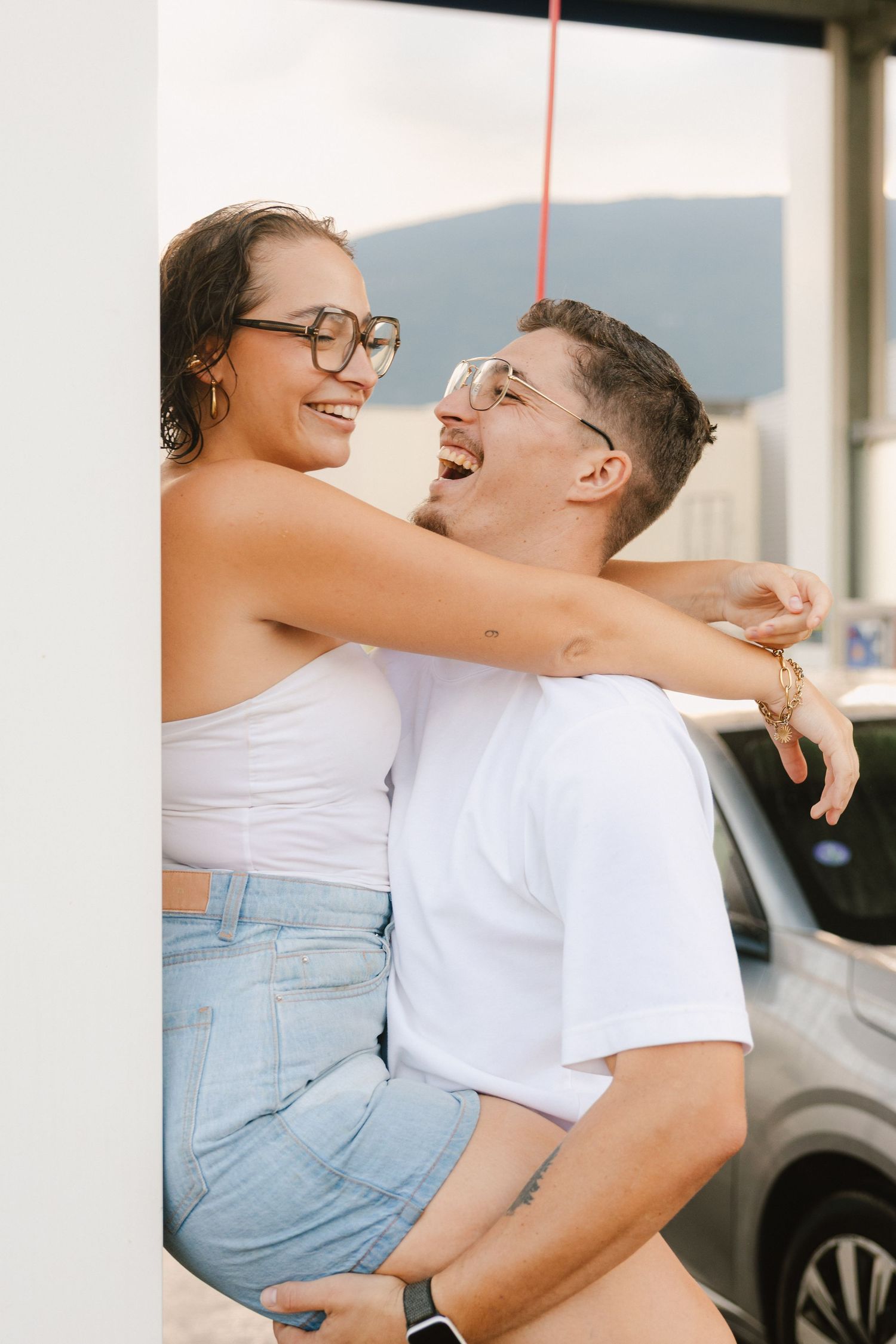 A romantic couple embraces outdoors while sharing a laugh together, dressed casually in white and denim.