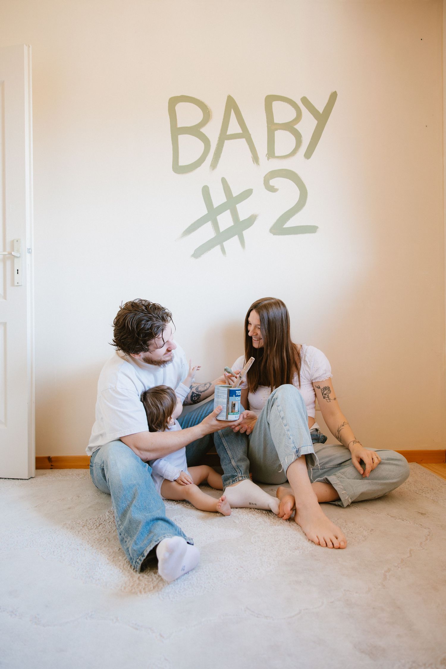A happy couple sits on the floor with their toddler, holding a photo, with Baby #2 written on the wall behind them.