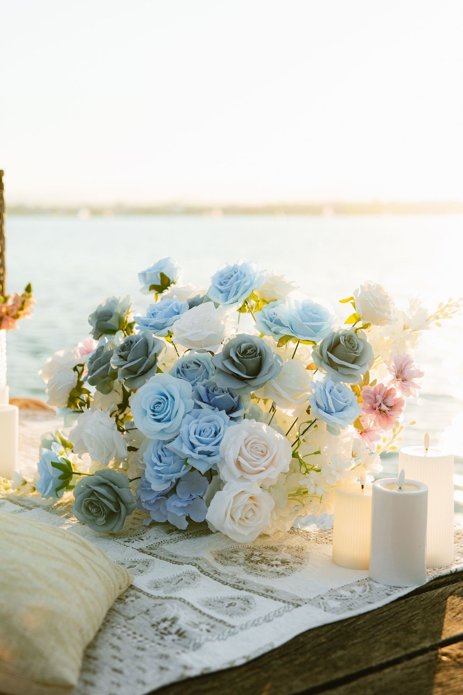 Romantic table setting with blue and white roses arranged on lace tablecloth with glowing candles creating soft ambiance.