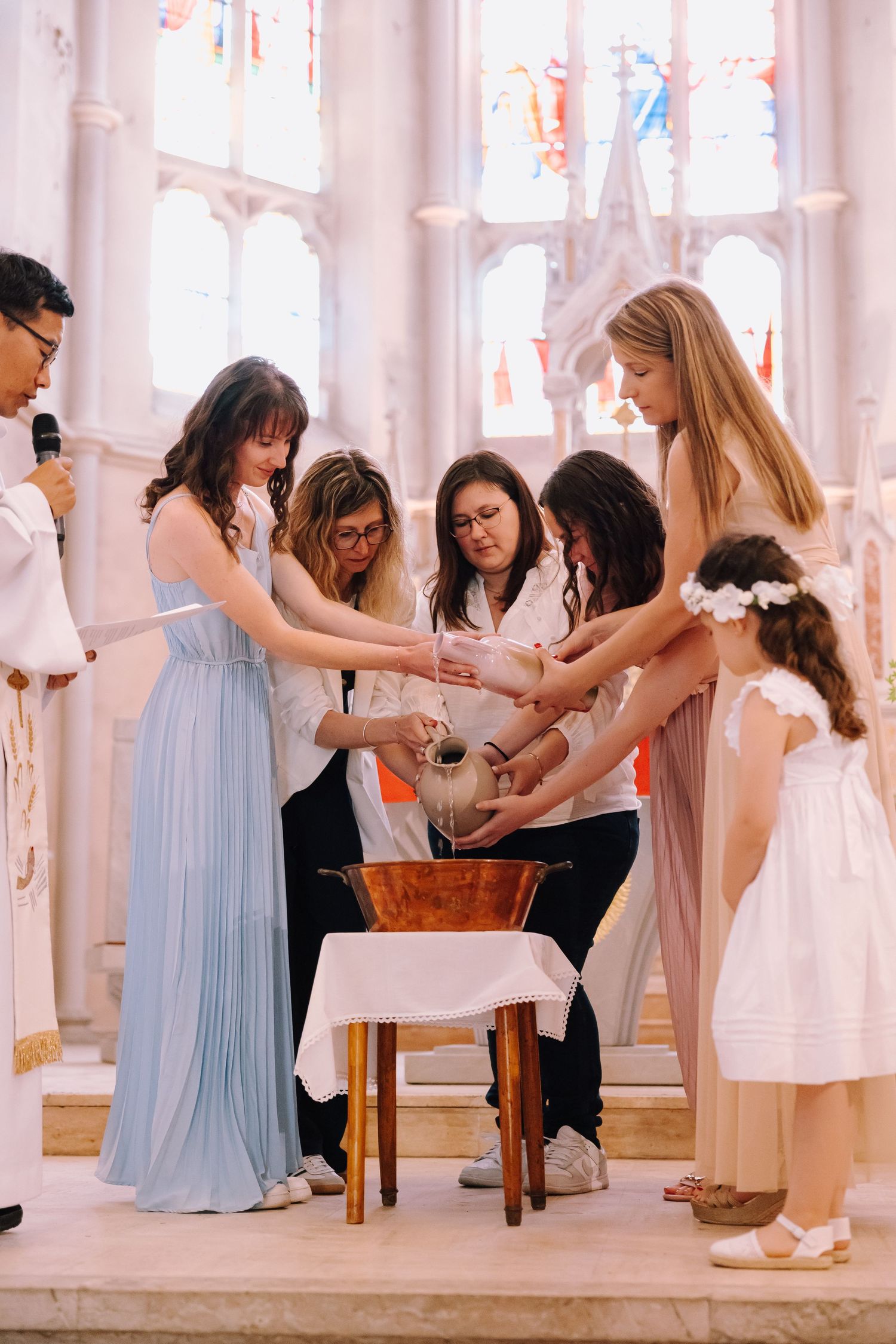 A group in light pastel colored dresses gathers around a small table in an ornate room with stained glass windows.