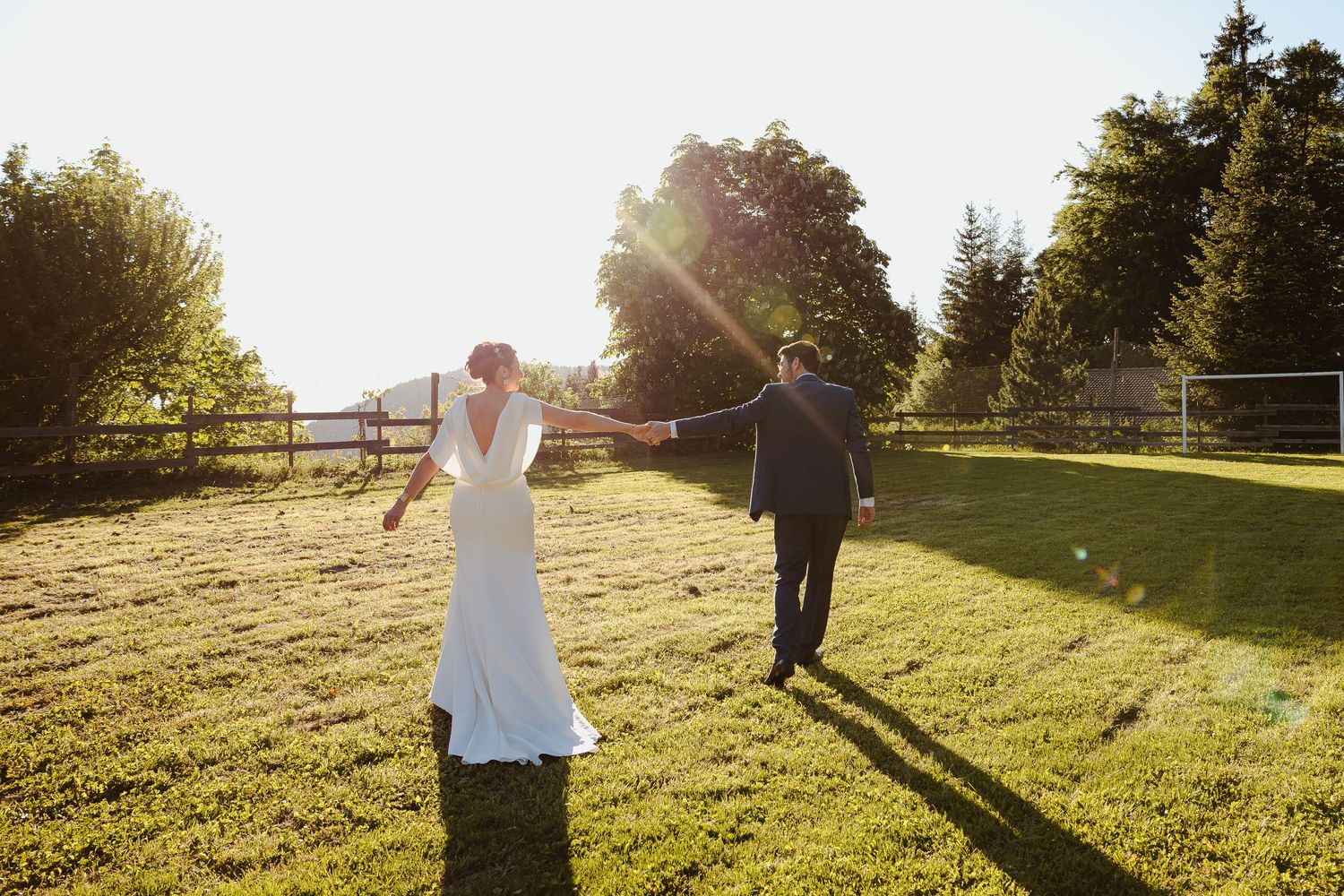 A couple in wedding attire walks through a sunlit meadow with a wooden fence and trees in the background.