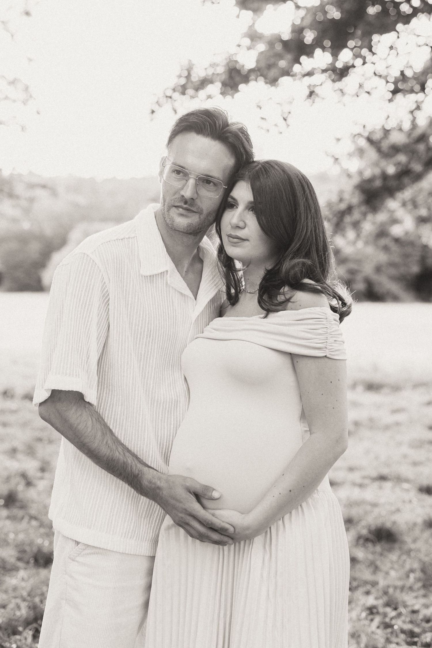 A black and white maternity photo of a couple embracing outdoors in white clothing during sunset.