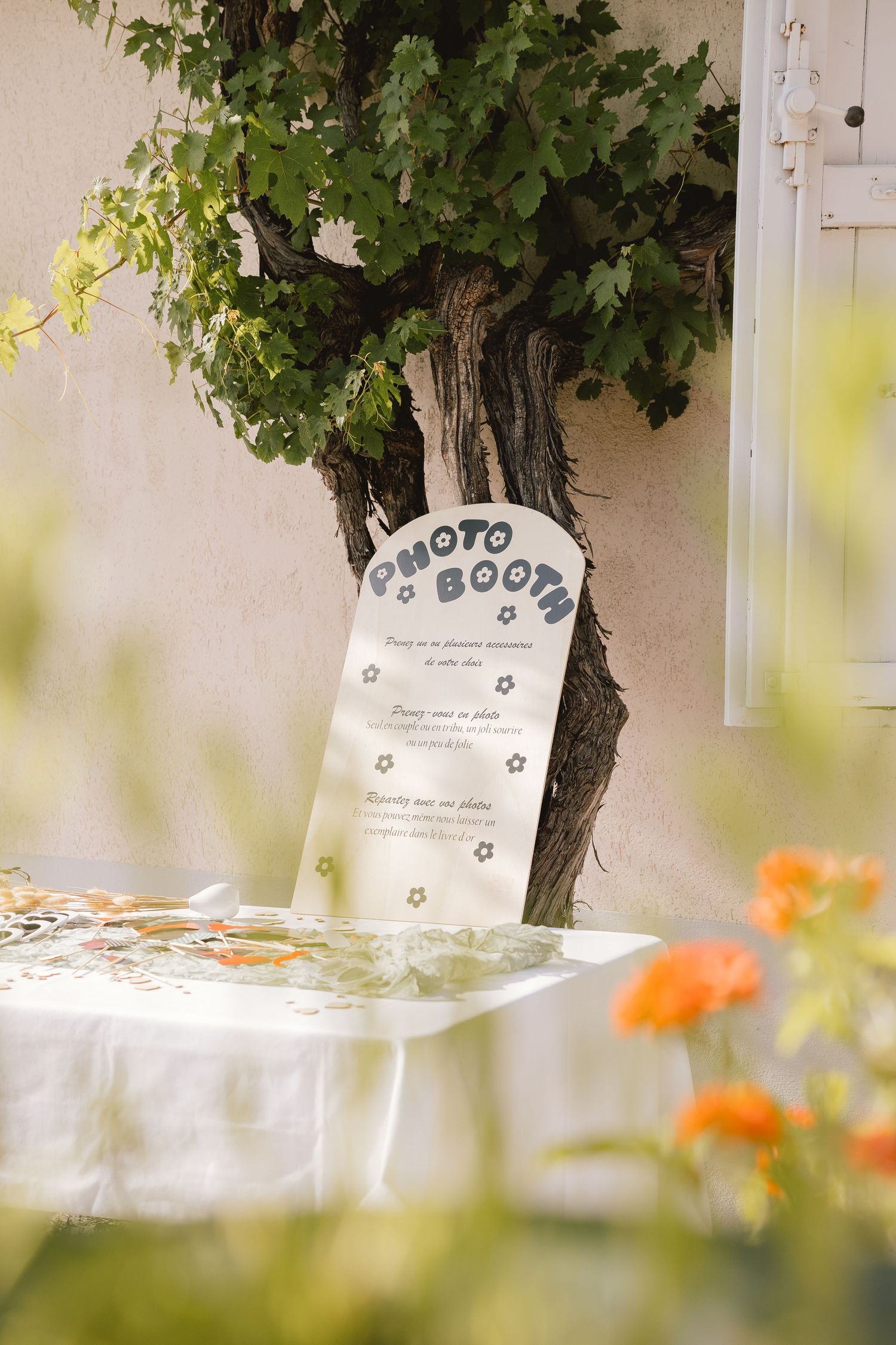 A photo booth sign stands against a grapevine-covered wall with orange flowers in the foreground at an outdoor event.