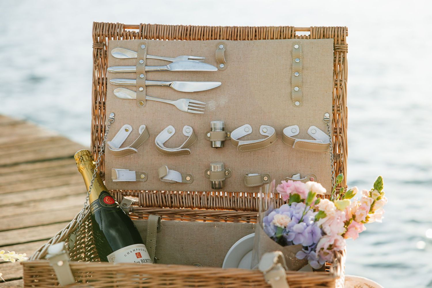 Rustic wicker picnic basket filled with elegant white dinnerware and wine glasses set against a beachy backdrop.