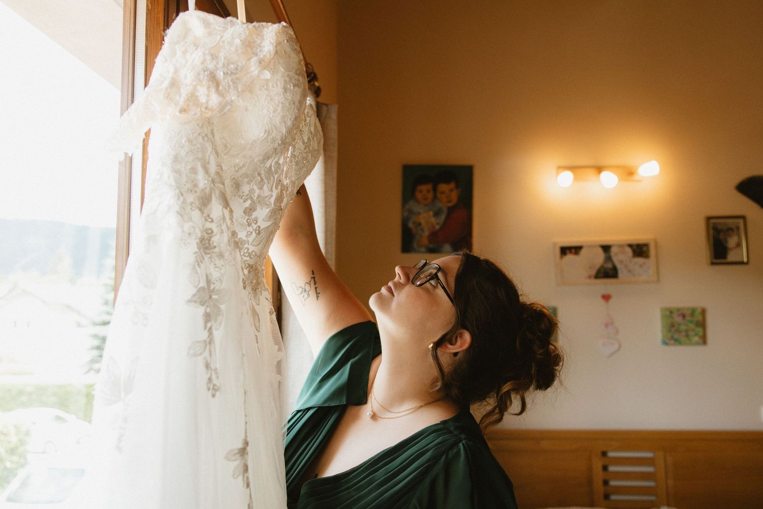A delicate lace wedding dress hangs elegantly against a warm-toned wall in natural lighting.