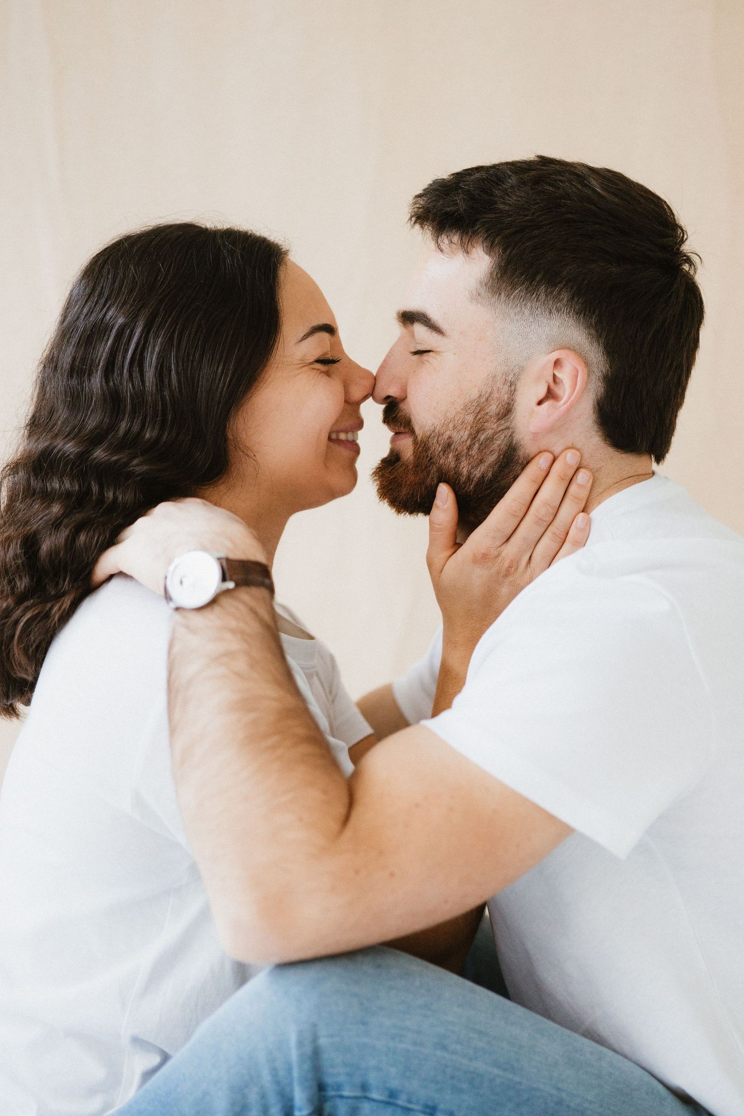 Intimate couple sharing tender moments while posing against beige wall in studio setting.