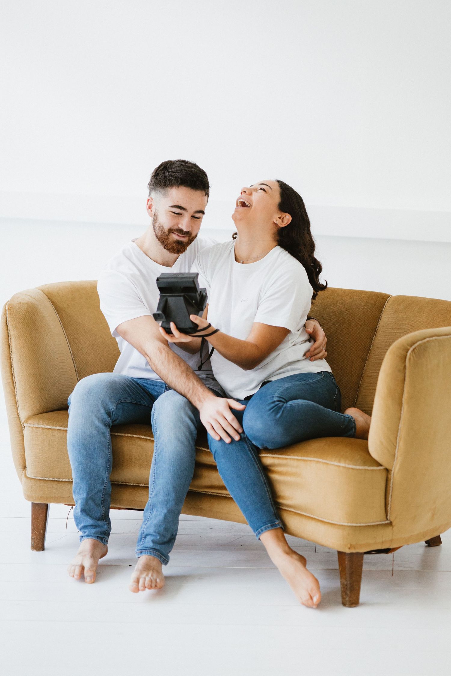 A couple shares a joyful moment together while sitting on tan armchairs in a bright minimalist setting.