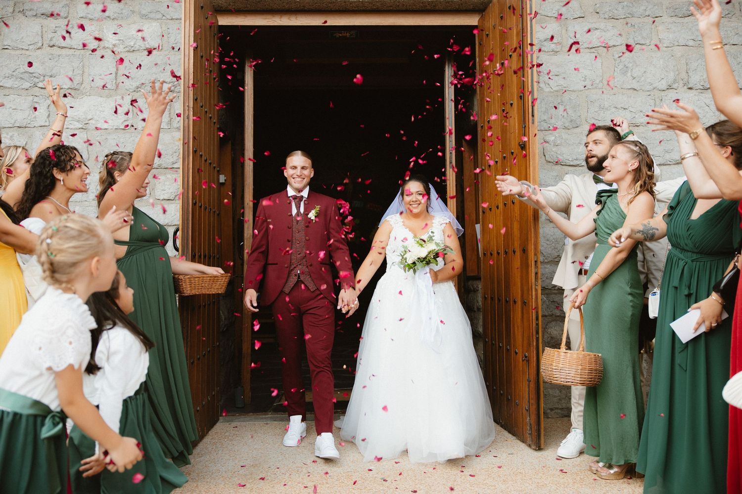 Wedding party celebrating with red flower petals being tossed in the air outside a rustic wooden building.