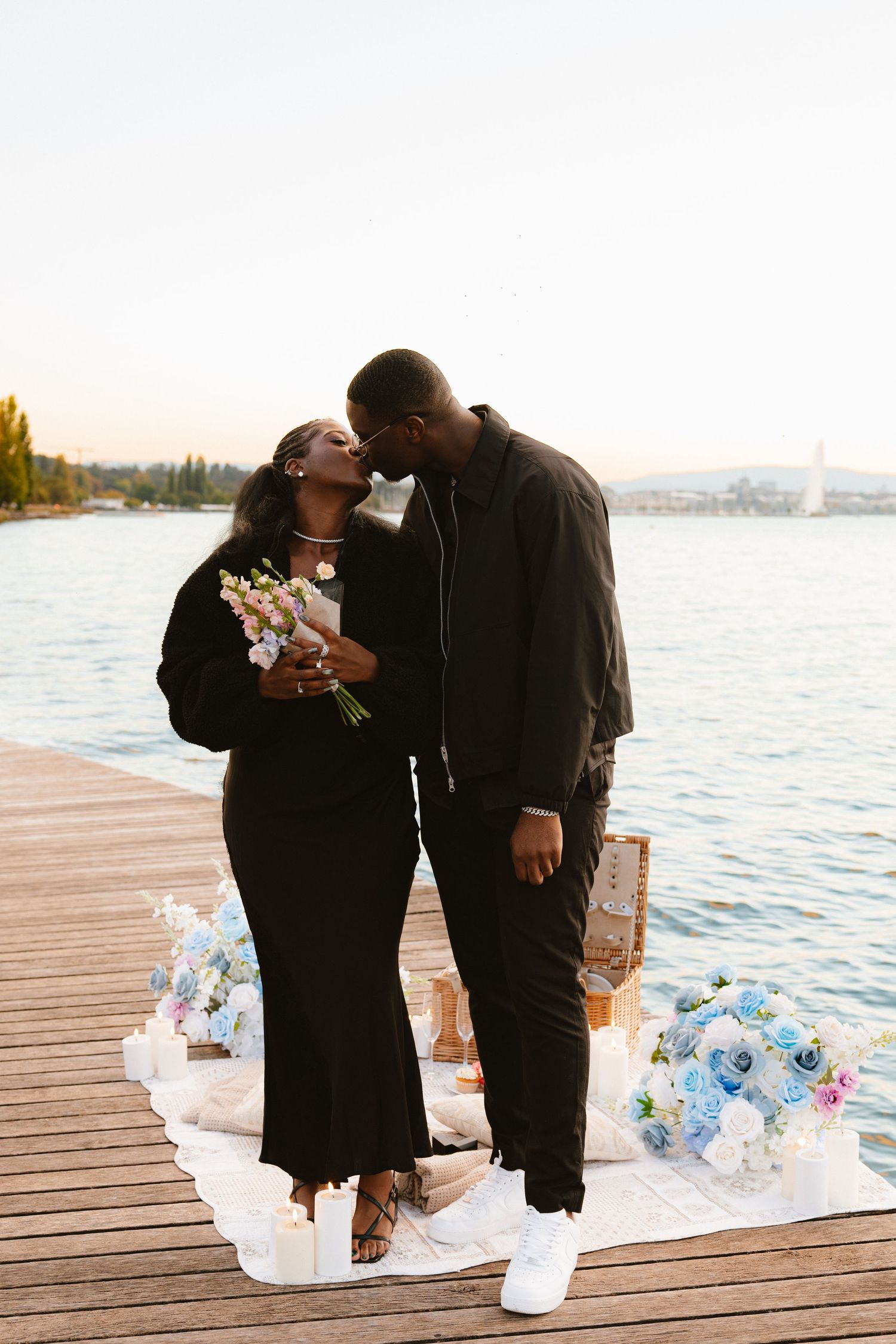 A romantic couple shares a tender moment on a wooden dock by the water surrounded by decorative bottles and flowers.