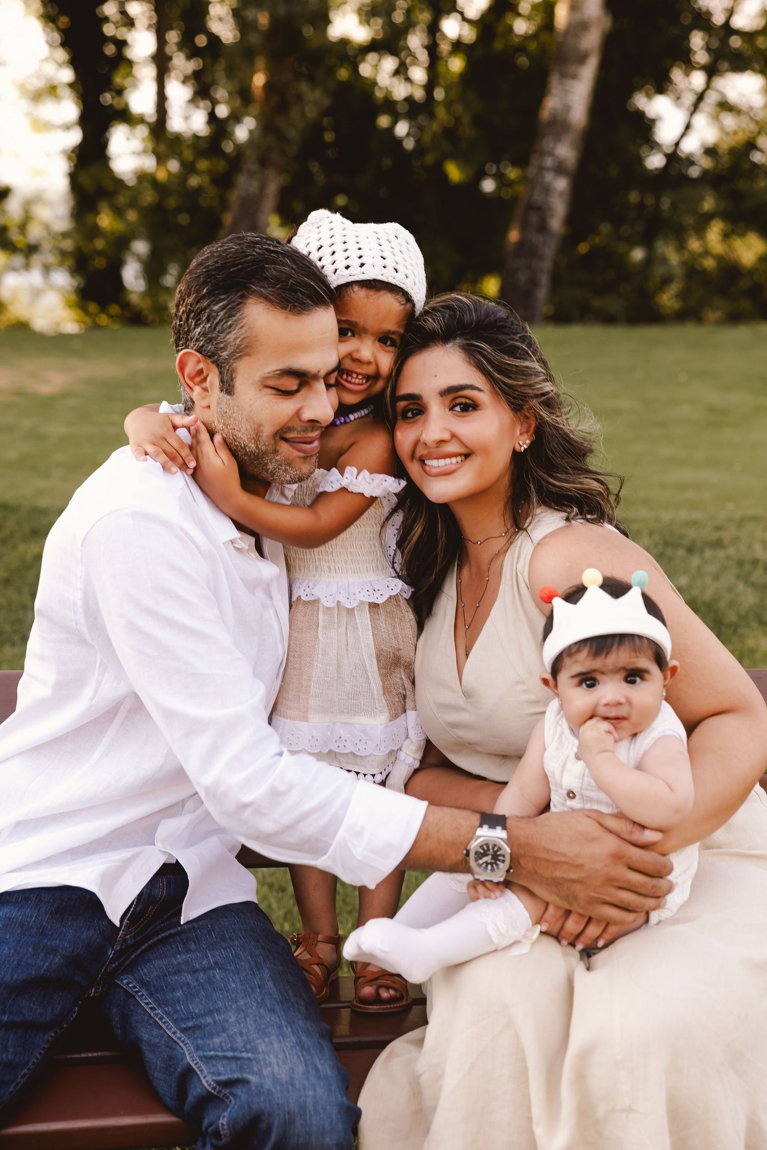 Happy family wearing white outfits sits close together on grass during golden hour outdoor photo session.