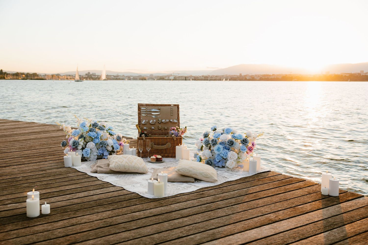 A romantic lakeside picnic setup with blue flowers, candles, and vintage decor on a wooden dock at sunset.