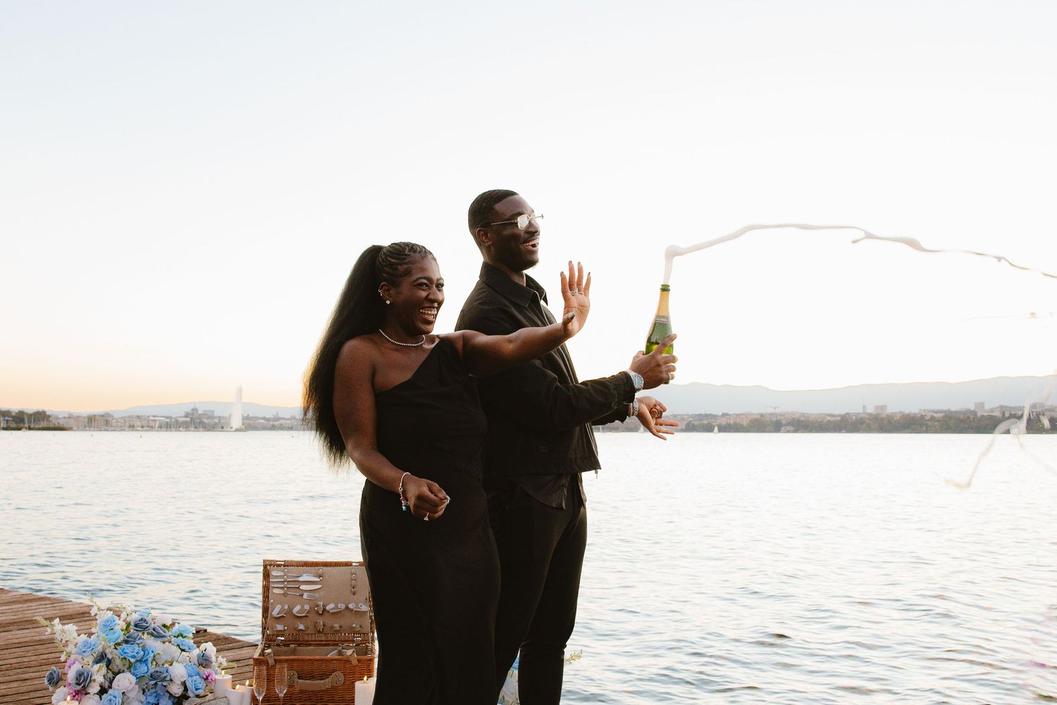 A couple celebrates by the water at sunset, popping champagne with a wooden crate nearby.
