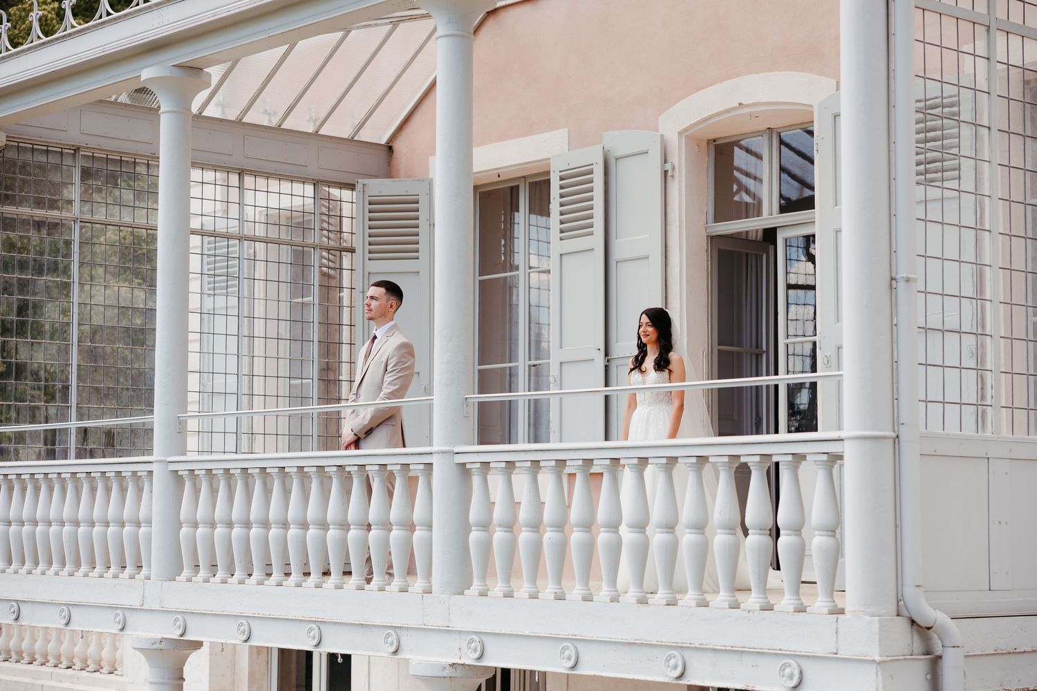 Two people in formal attire stand separately on an elegant white balcony with ornate railings and columns.