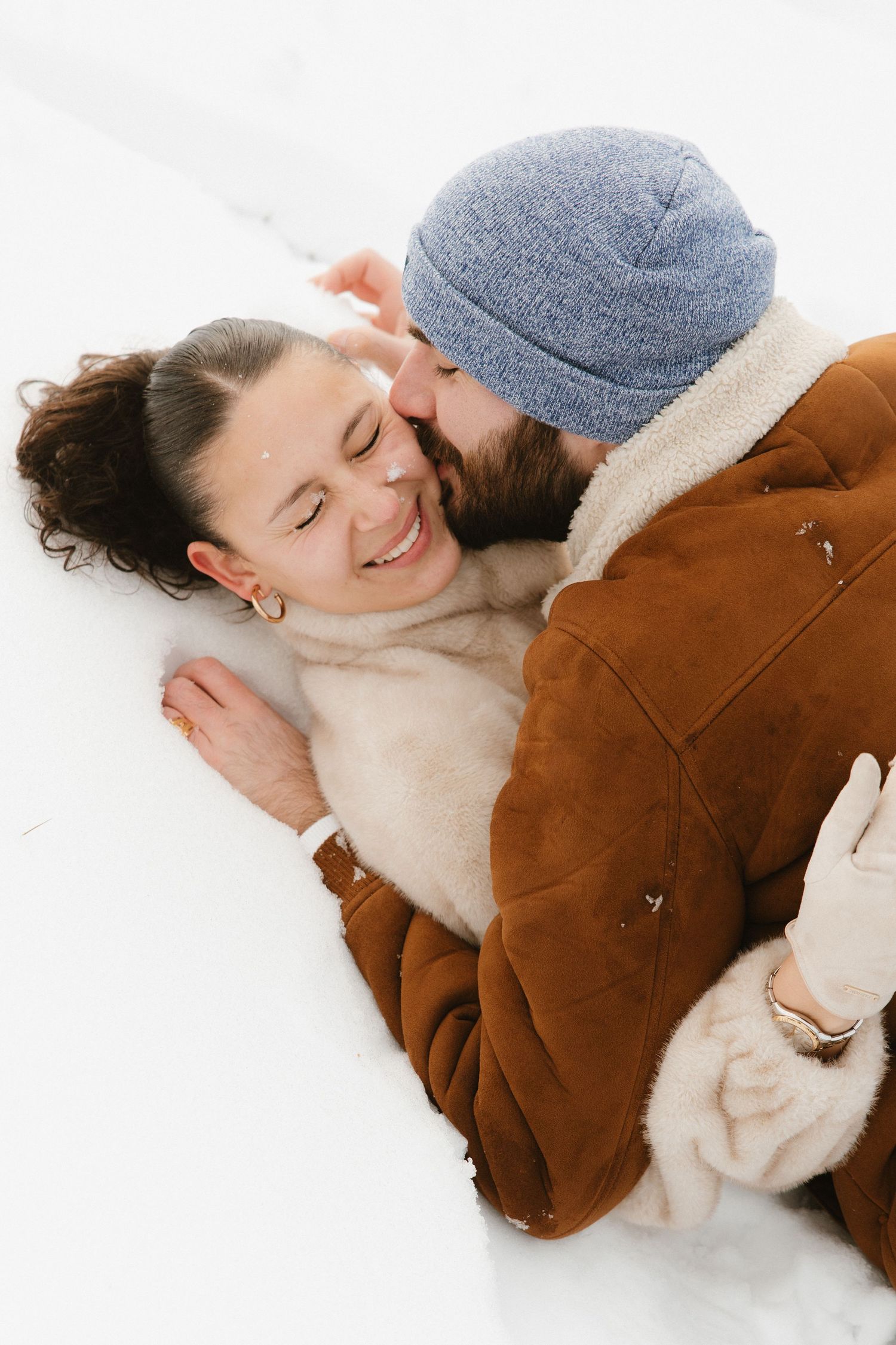 Person lying in snow wearing brown suede jacket and gray beanie while smiling with eyes closed.
