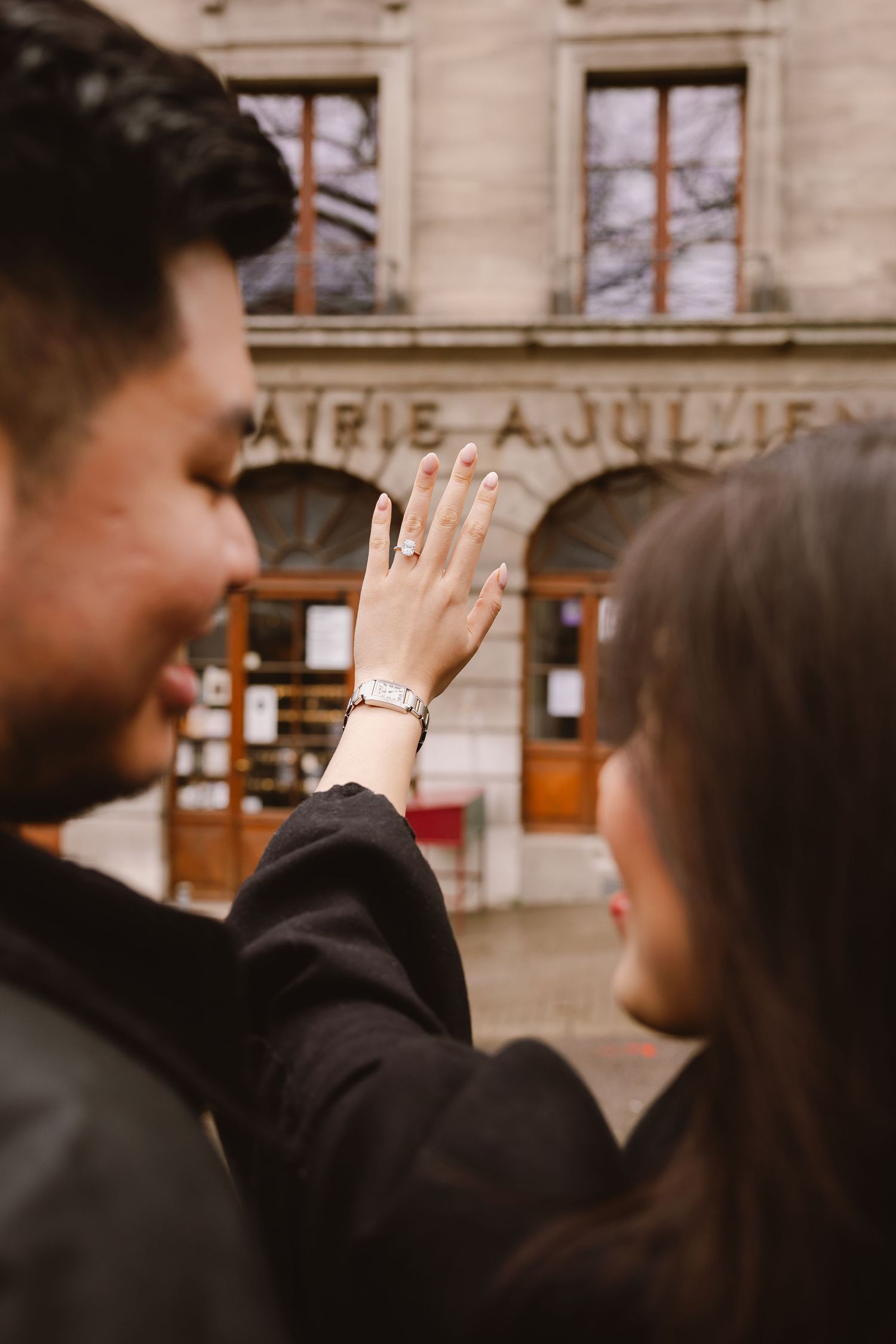 Two figures stand close together while one holds up their hand with an engagement ring on a fall day outside a building.