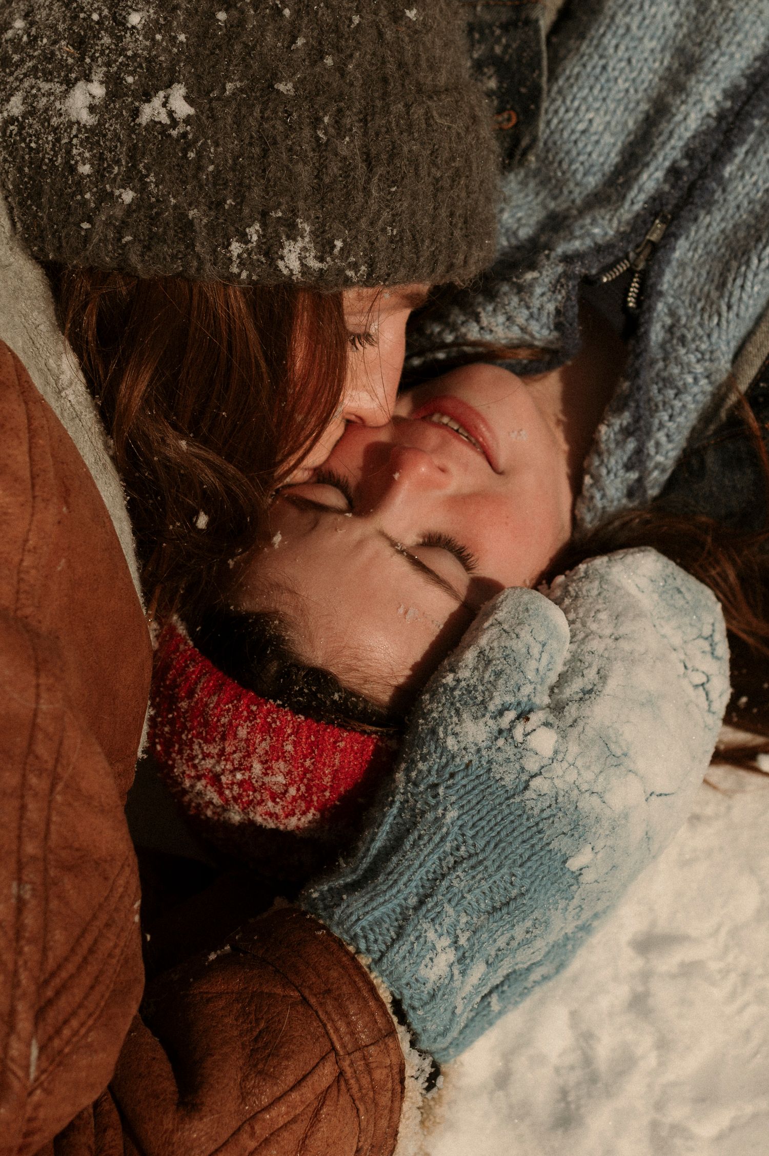mom kisses daughter while laying in the snow, wearing a hat and gloves