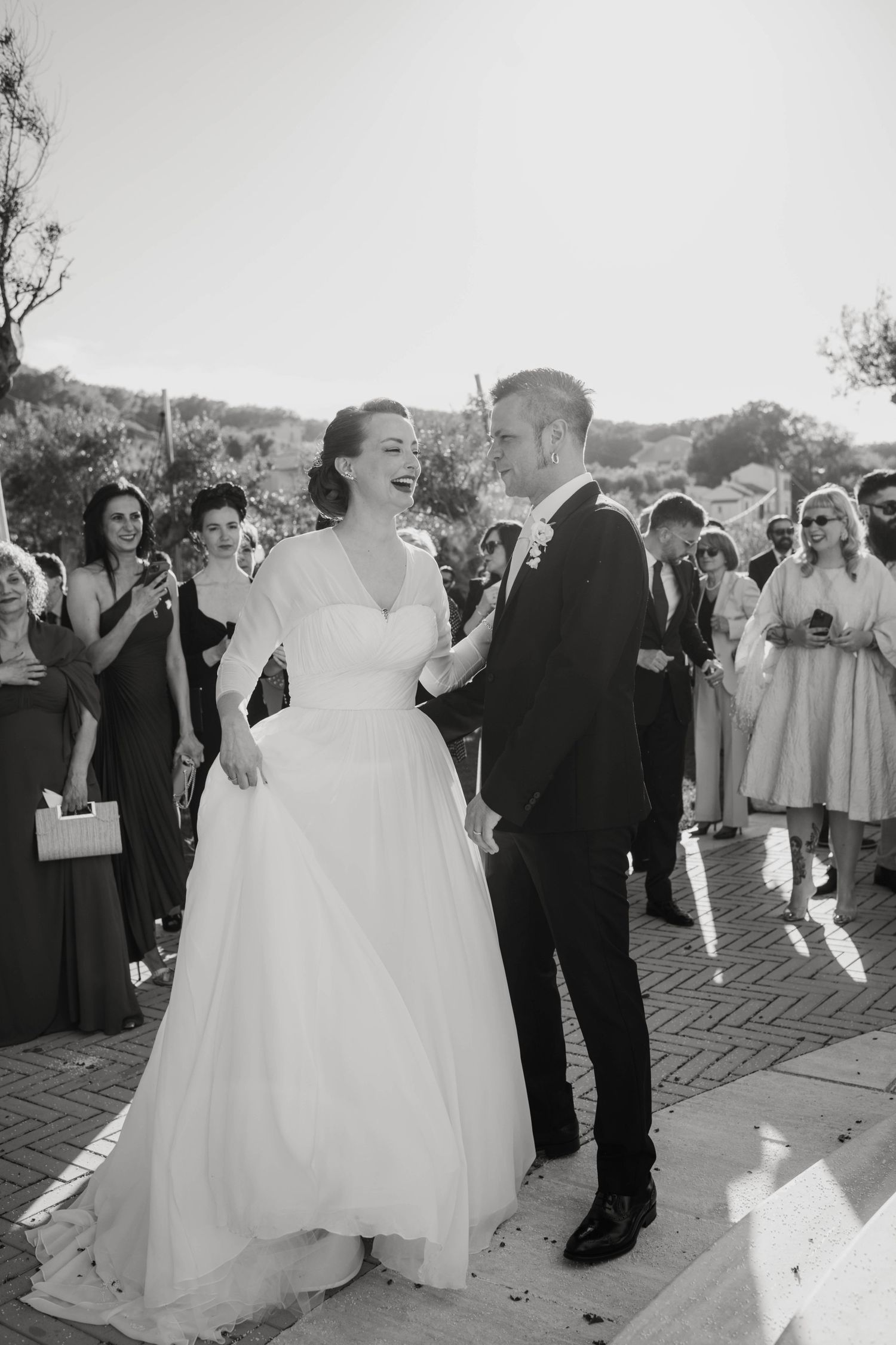 Black and white candid wedding photo showing a bride in a classic white gown laughing with guests at an outdoor ceremony.