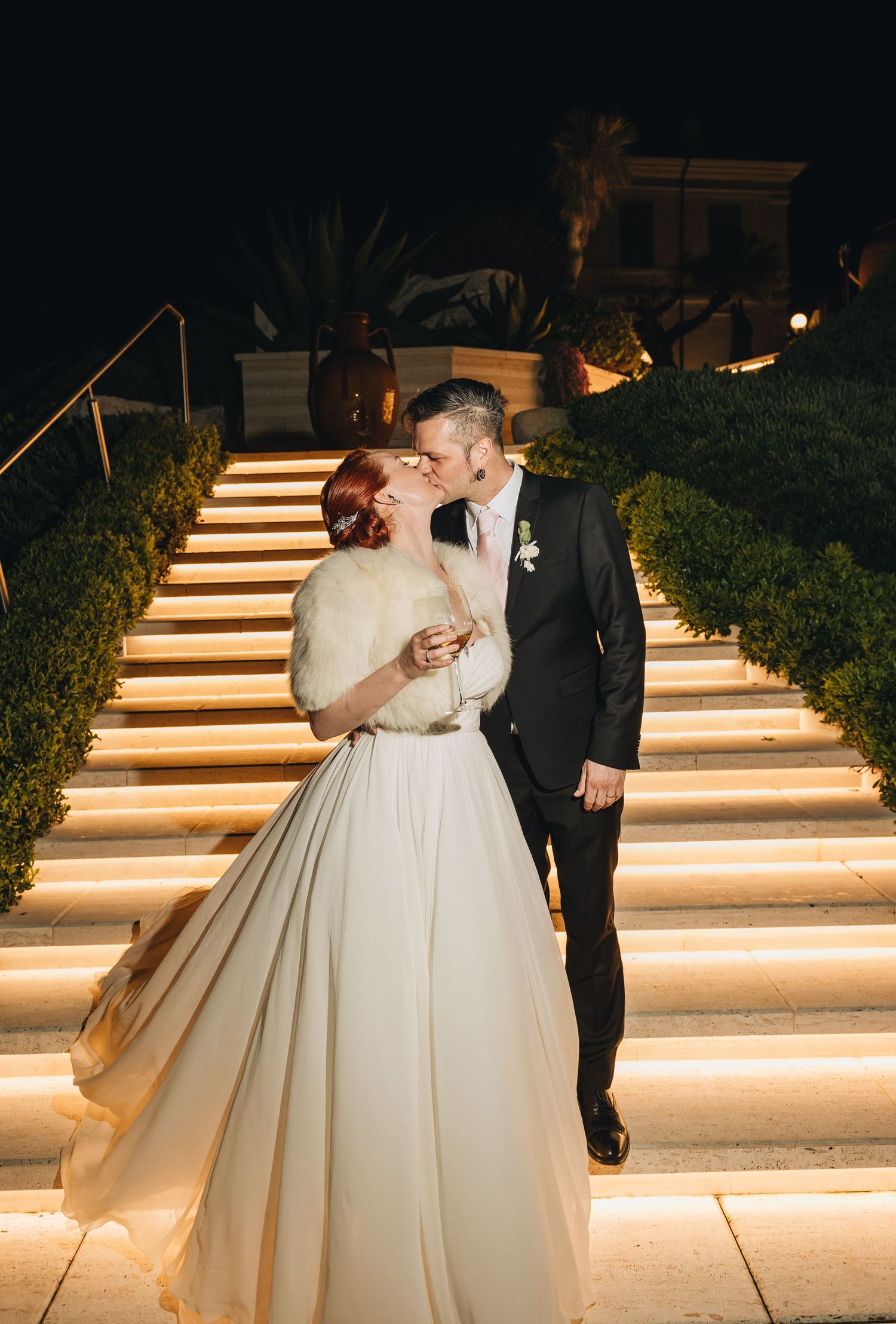 A couple share a romantic kiss on illuminated stone steps at night during their elegant outdoor wedding celebration.