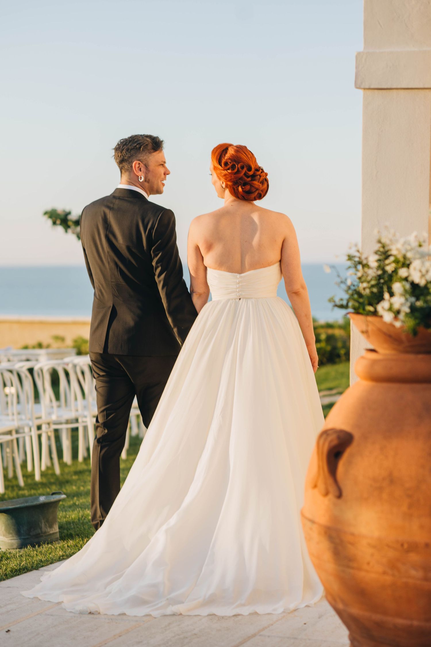 A bride in white gown and groom in dark suit gaze at ocean view from hilltop terrace with rustic pottery decor.