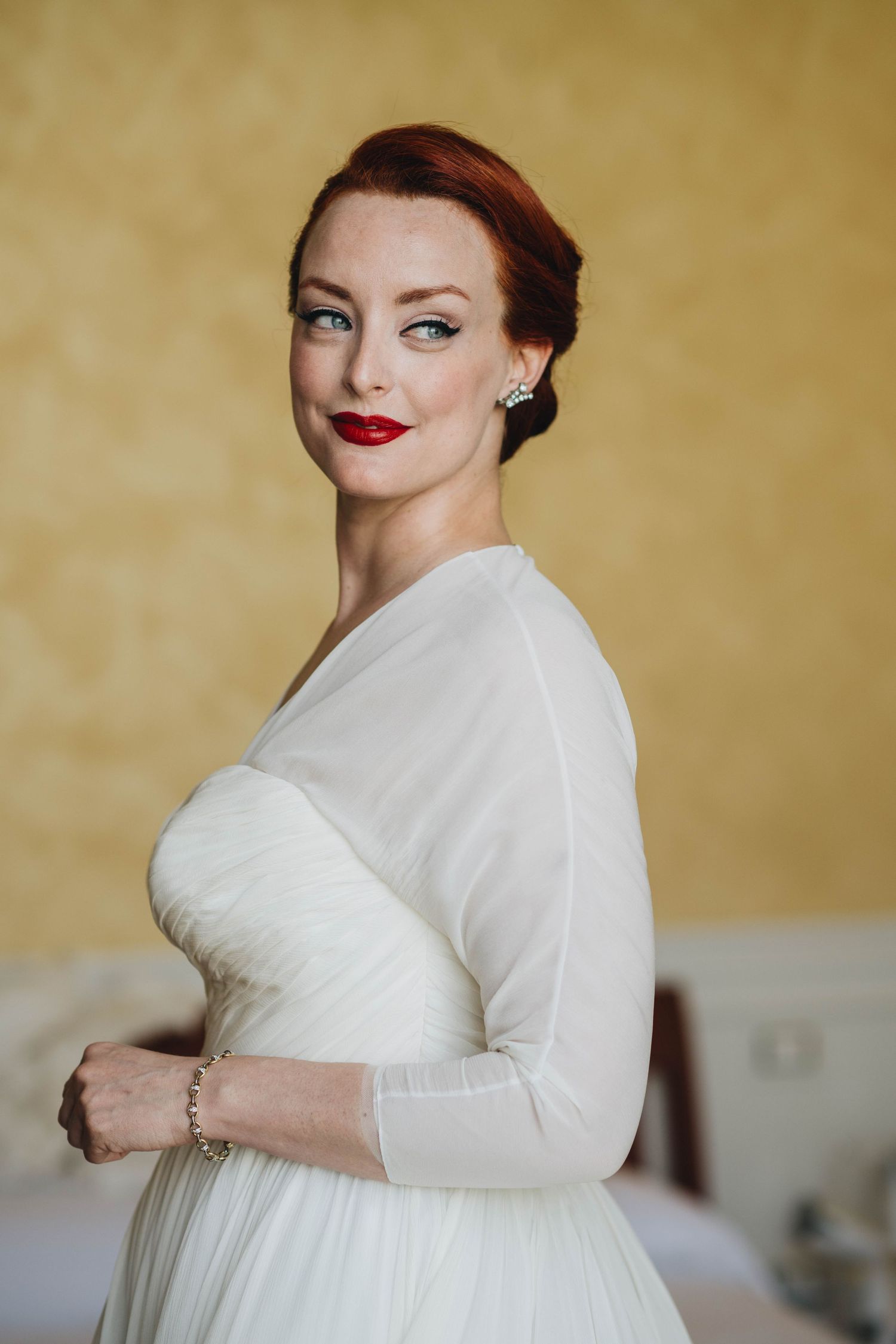 Elegant woman in white long-sleeved wedding dress with vintage-inspired makeup and upswept hairstyle.