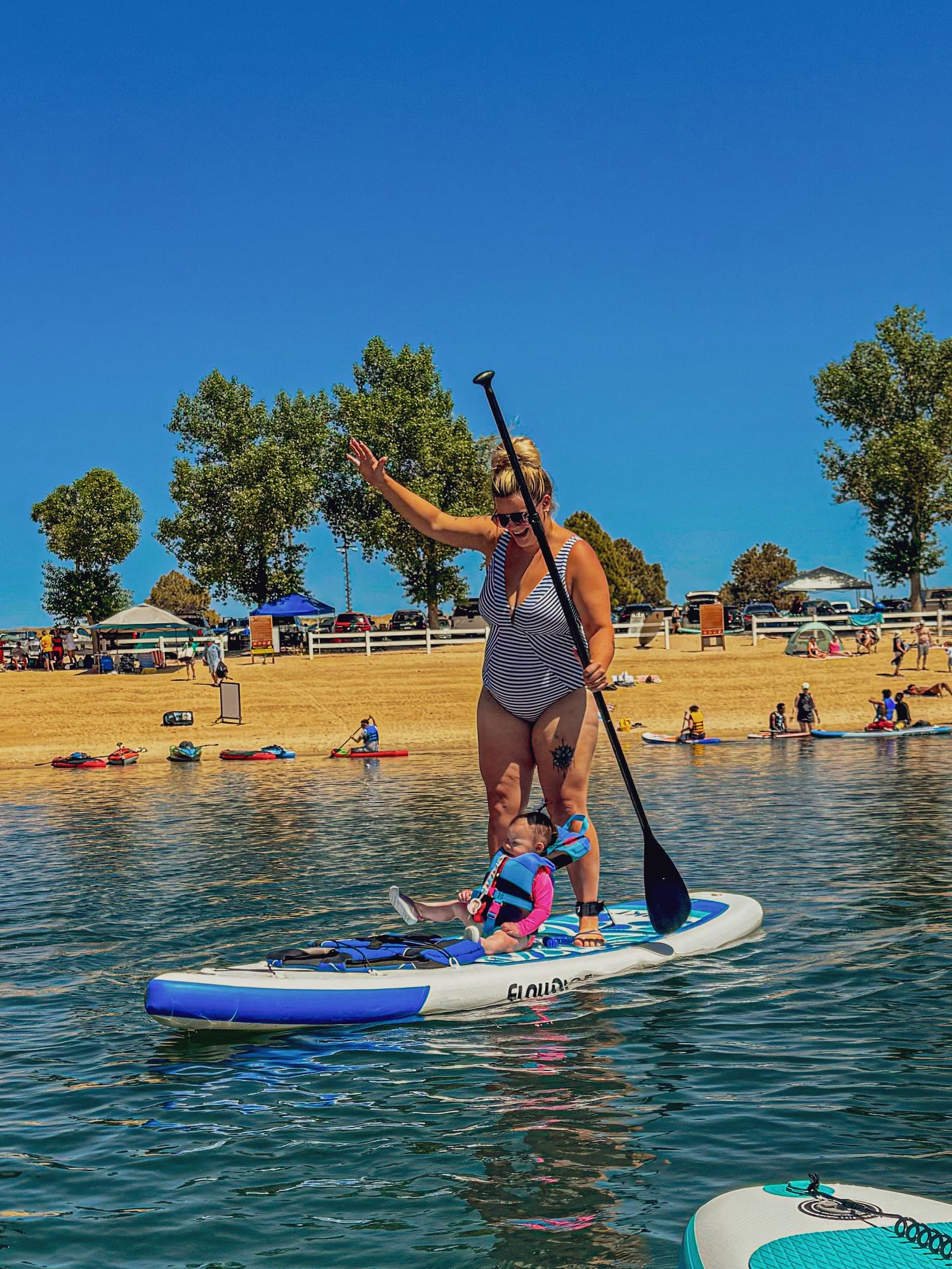 Person paddleboarding on a lake during a sunny summer day with beachgoers in background.