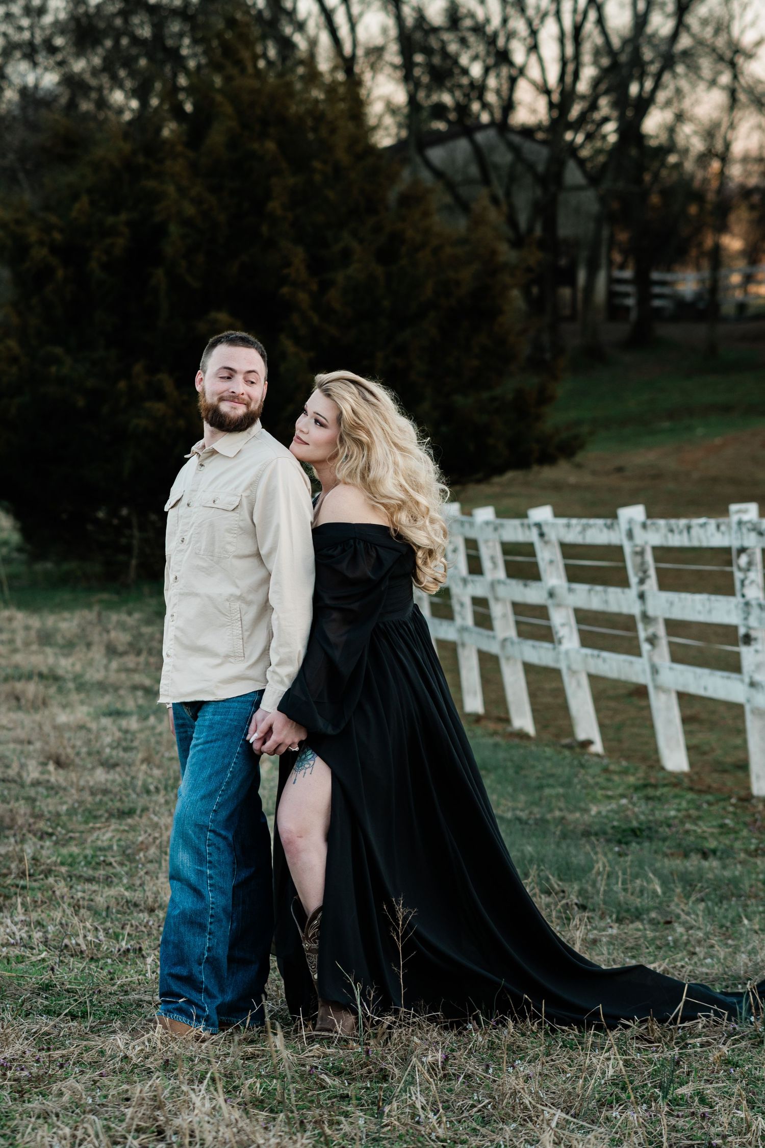 Couple in contrasting black and white outfits posing near white fence in countryside setting.