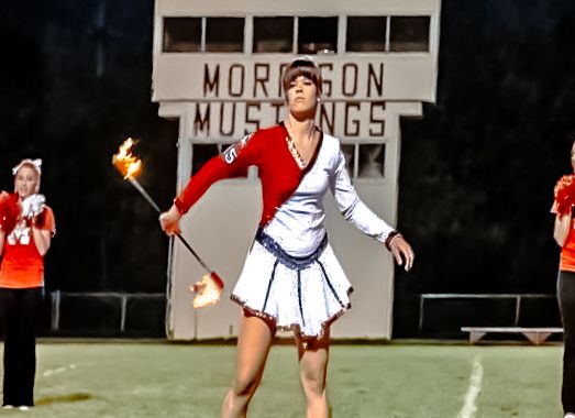 Baton twirler in red and white uniform performs with fire on field at Morrison Mustangs stadium.