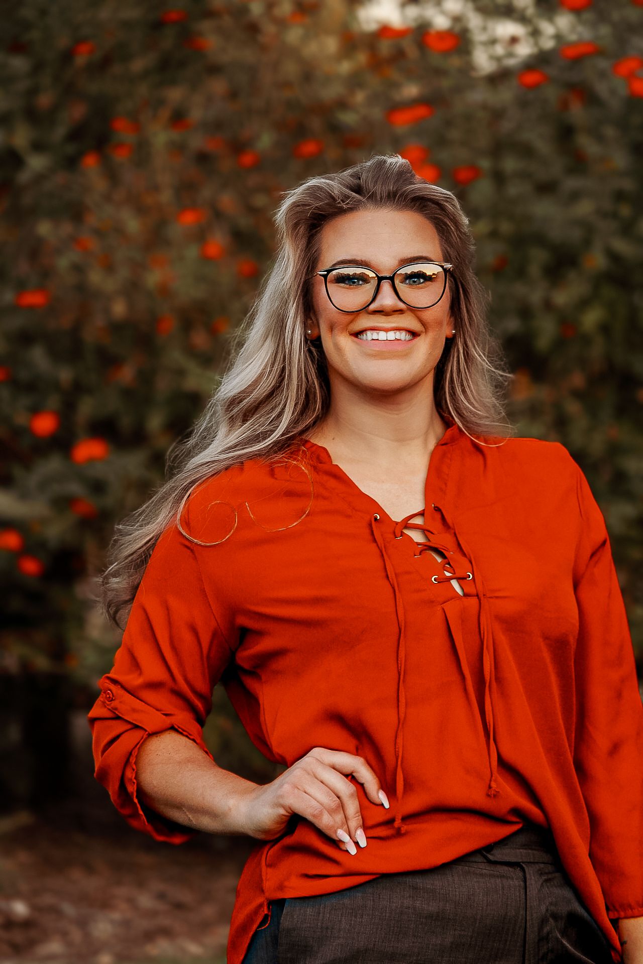 Woman in bright red blouse posing outdoors with autumn foliage in background.