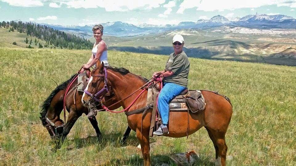 Two riders on horseback pause in a scenic mountain meadow with snow-capped peaks in the distance.