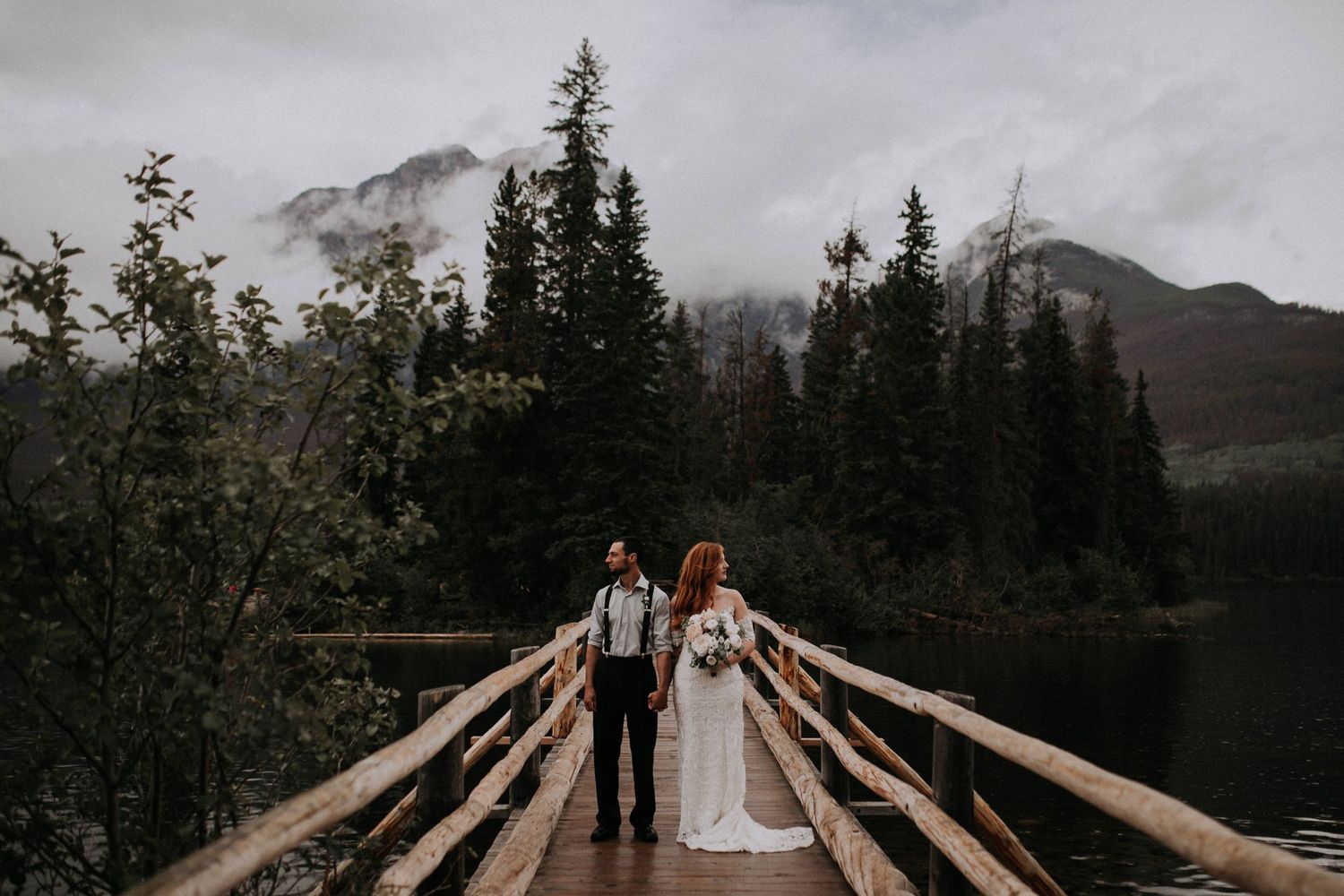 Two people stand on a wooden bridge over a lake surrounded by misty mountains and evergreen trees in the wilderness.