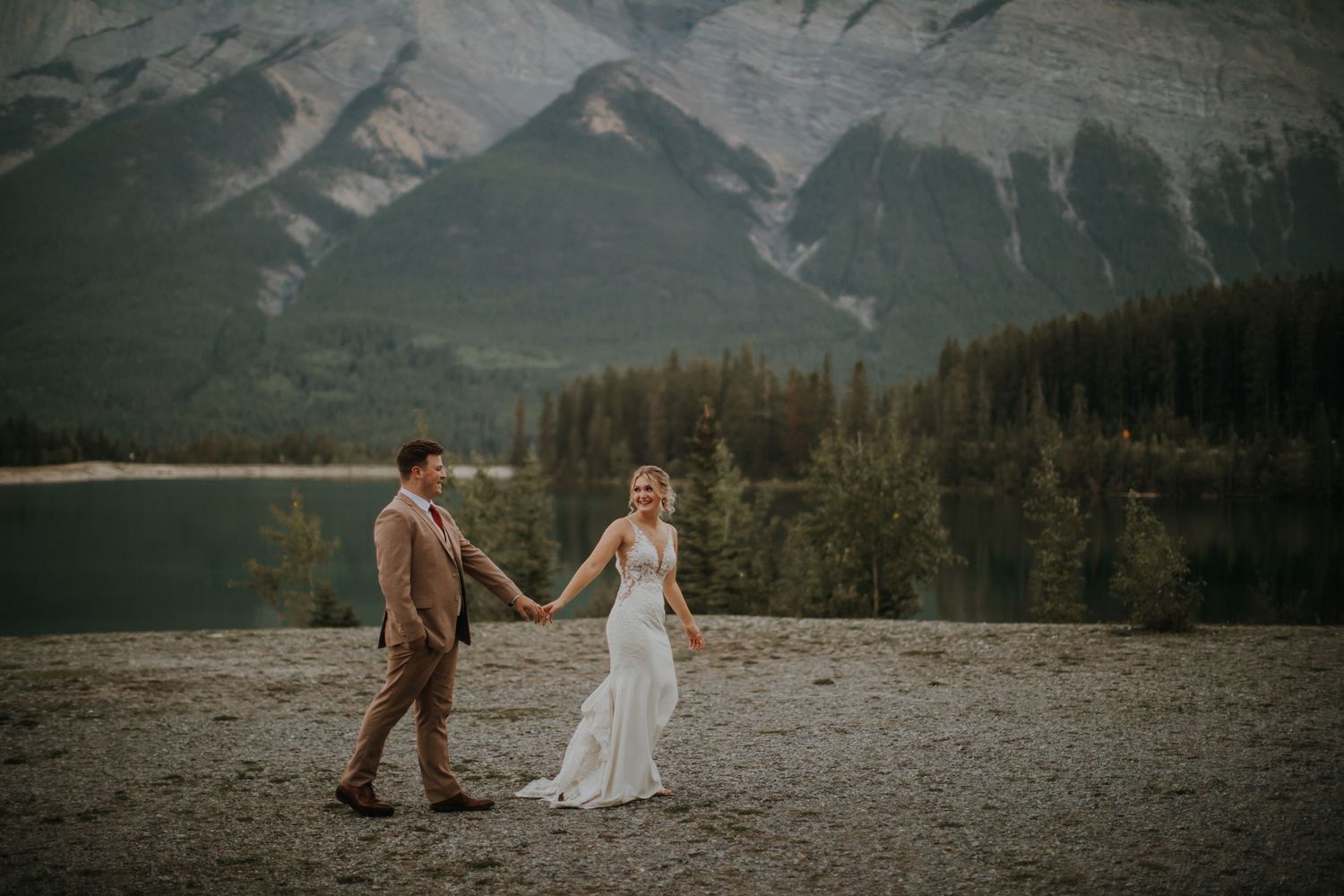 A couple in wedding attire holds hands against a dramatic mountain backdrop at a lake in Banff National Park.