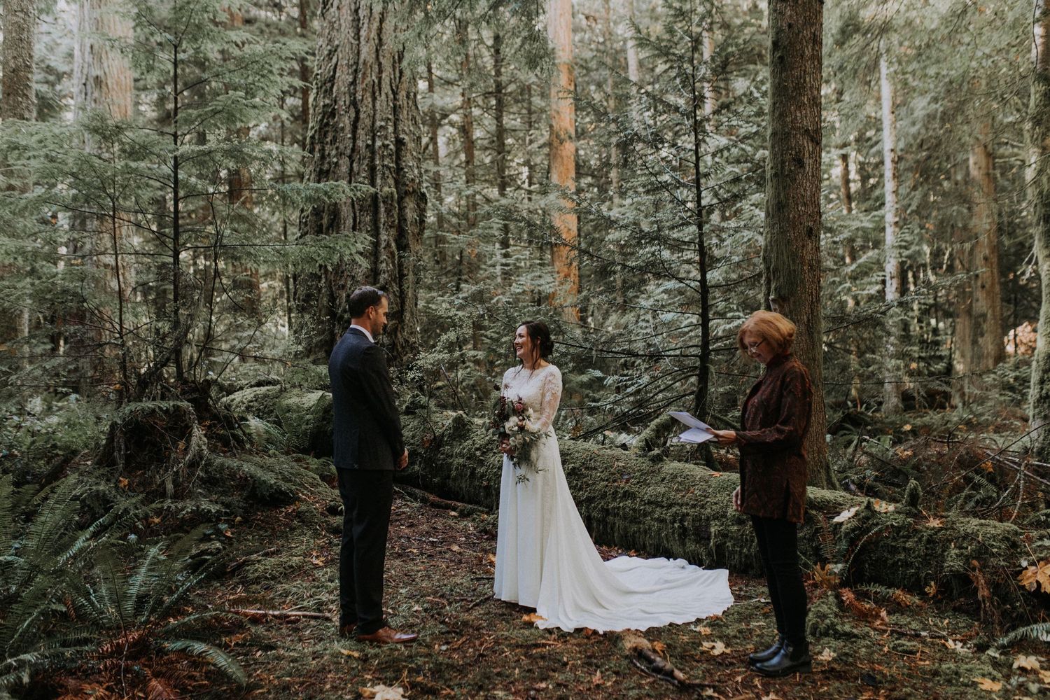 An intimate forest wedding ceremony takes place among tall evergreen trees and ferns in a misty woodland setting.