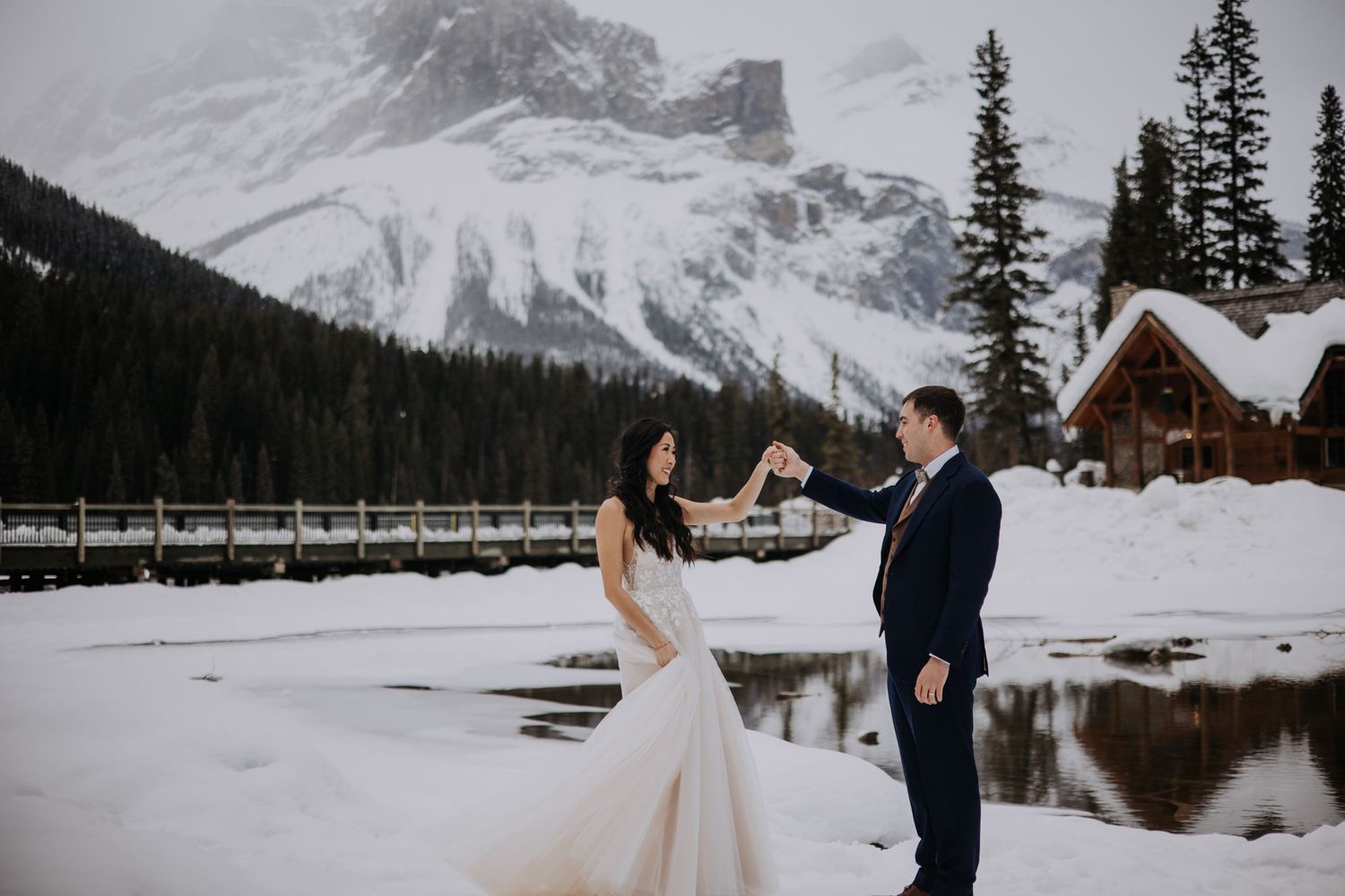 Couple in wedding attire sharing a moment by snowy Emerald Lake with mountain peaks and rustic lodge in background.