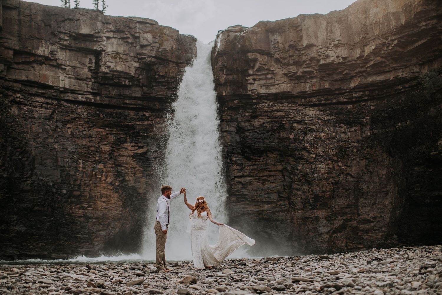 A wedding couple poses dramatically in front of a majestic waterfall against rocky cliffs.