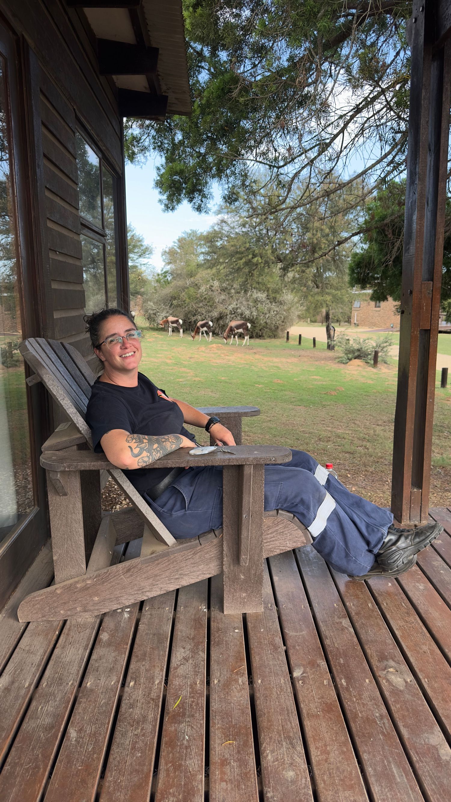 Person relaxing in wooden Adirondack chair on rustic porch with green lawn and trees in background.