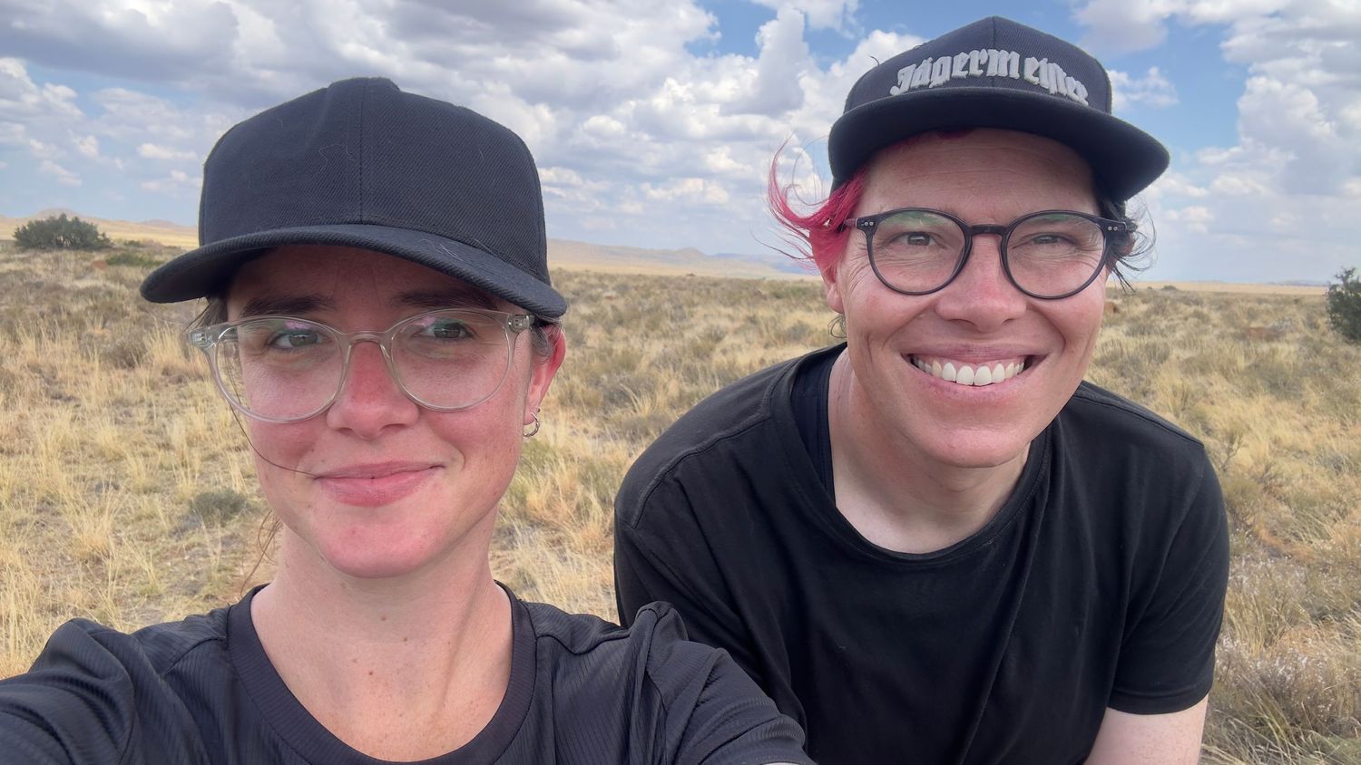Two smiling people wearing baseball caps take a selfie outdoors in an arid grassland landscape.