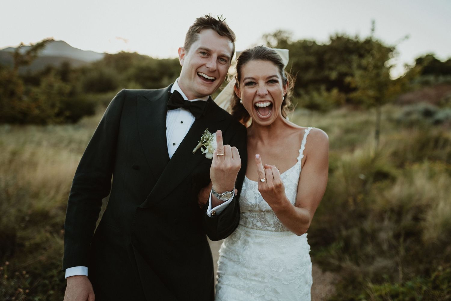 An excited couple showing off their rings in formal attire against a natural landscape.