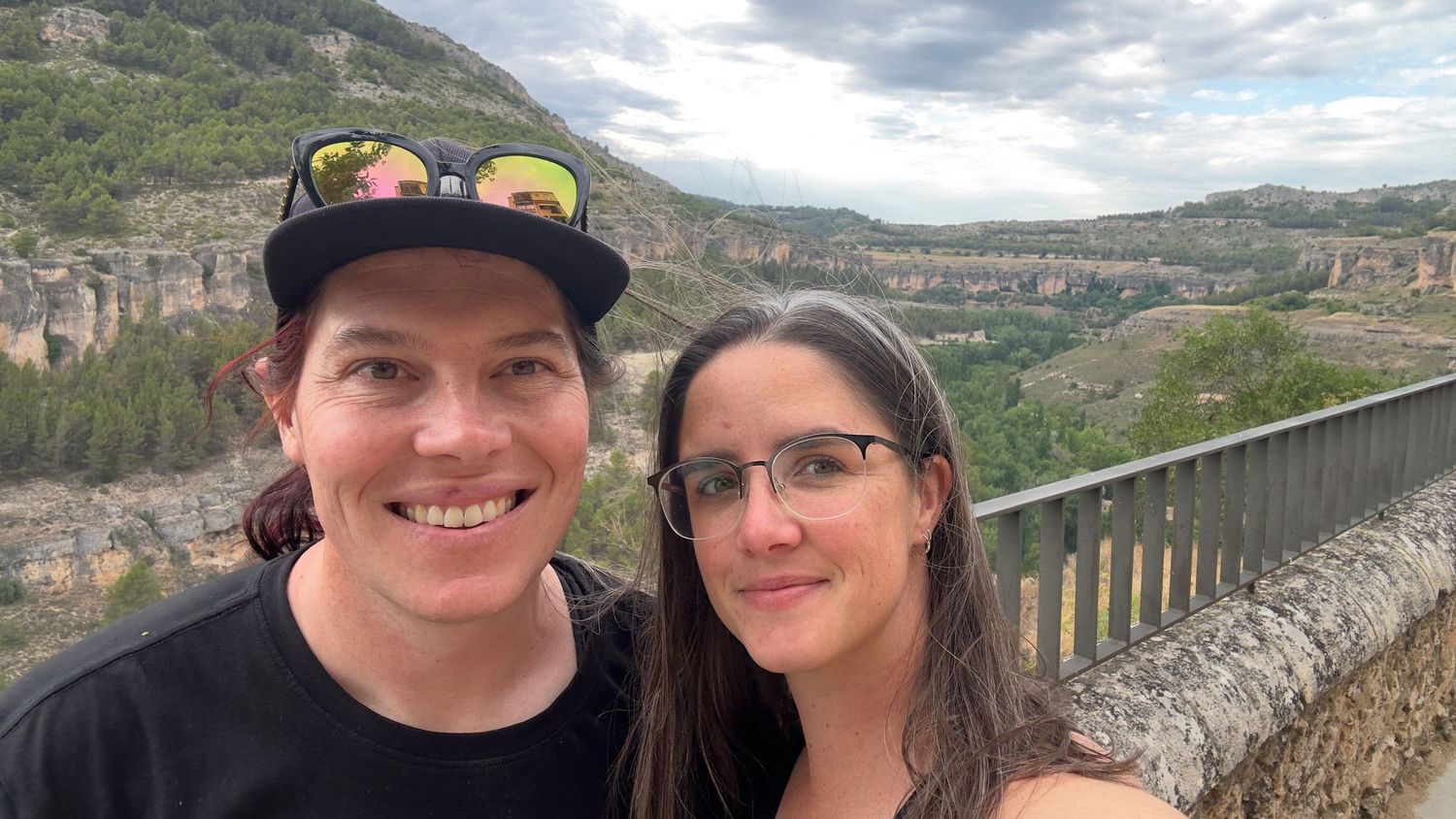 A smiling couple poses for a selfie on a stone bridge overlooking a lush green canyon landscape.