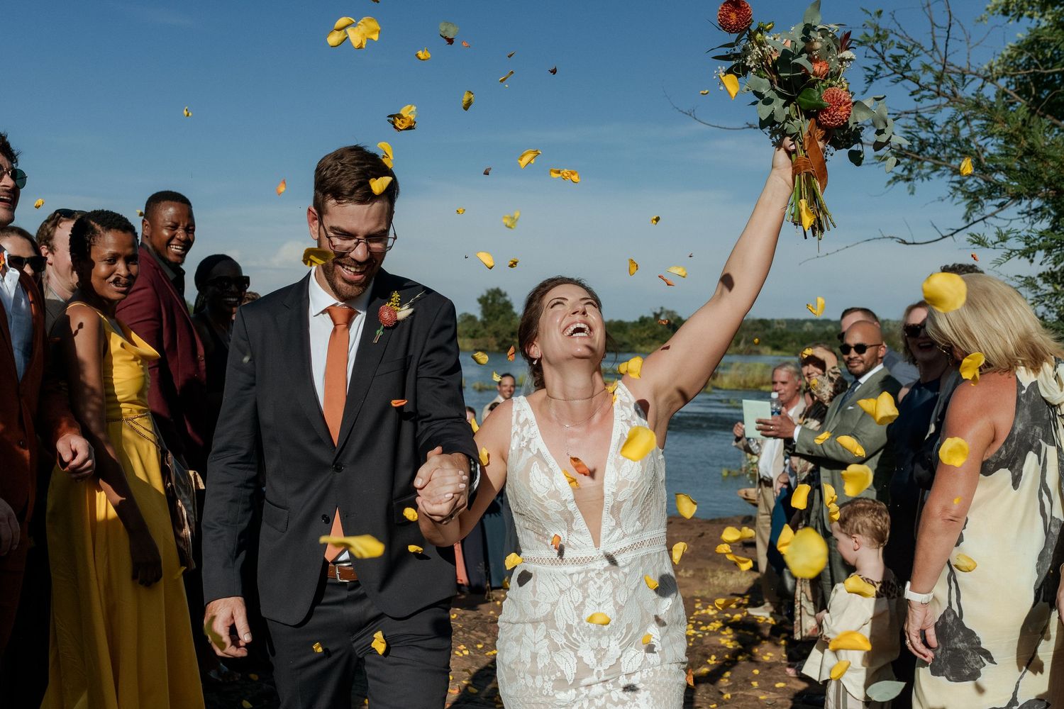 Wedding party celebrates outdoors with yellow flower petals being tossed in the air during a joyful moment.