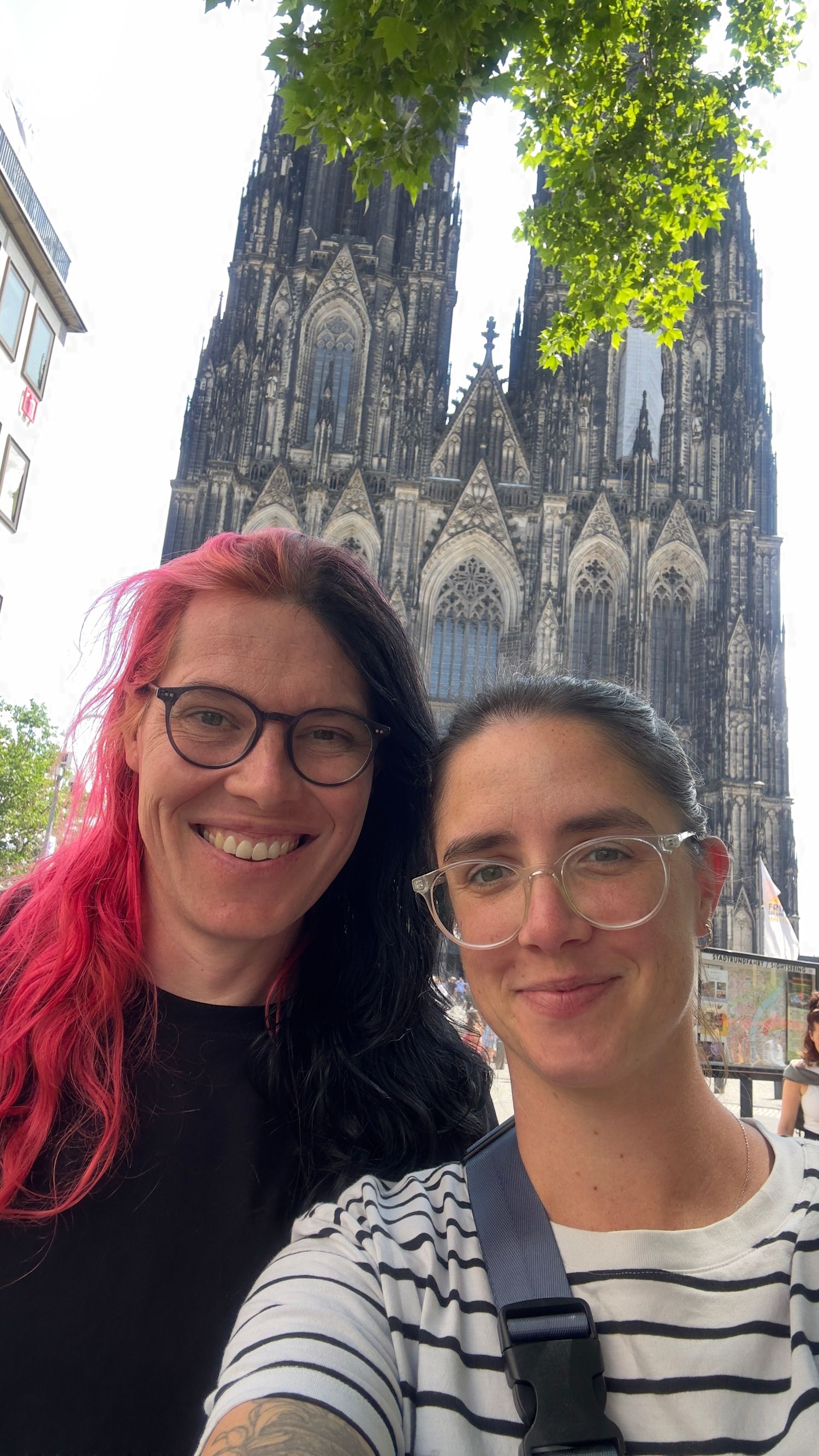 Two smiling women take a selfie in front of the iconic Cologne Cathedral in Germany on a sunny day.
