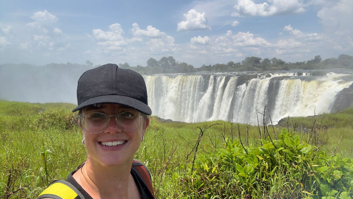 Woman smiling in selfie with Victoria Falls waterfall and lush greenery in background on sunny day.