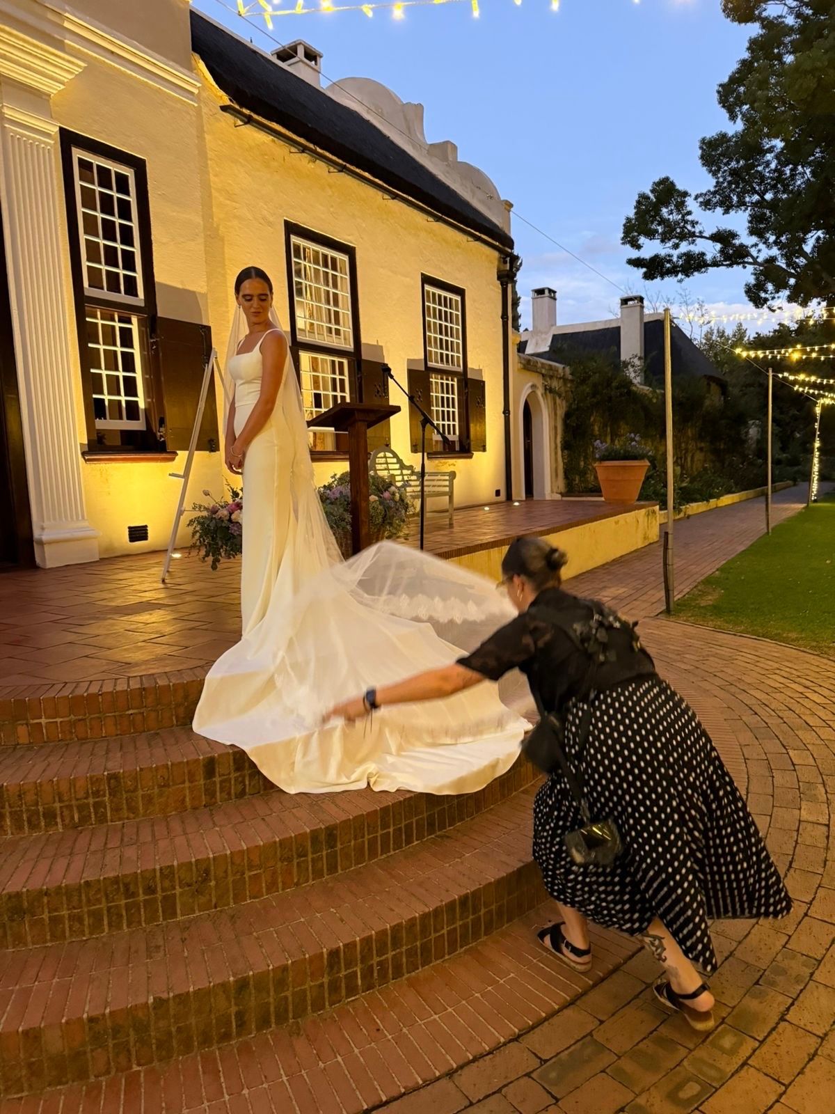 A bride in a white gown stands on brick steps while an assistant arranges her train outside an elegant building at dusk.