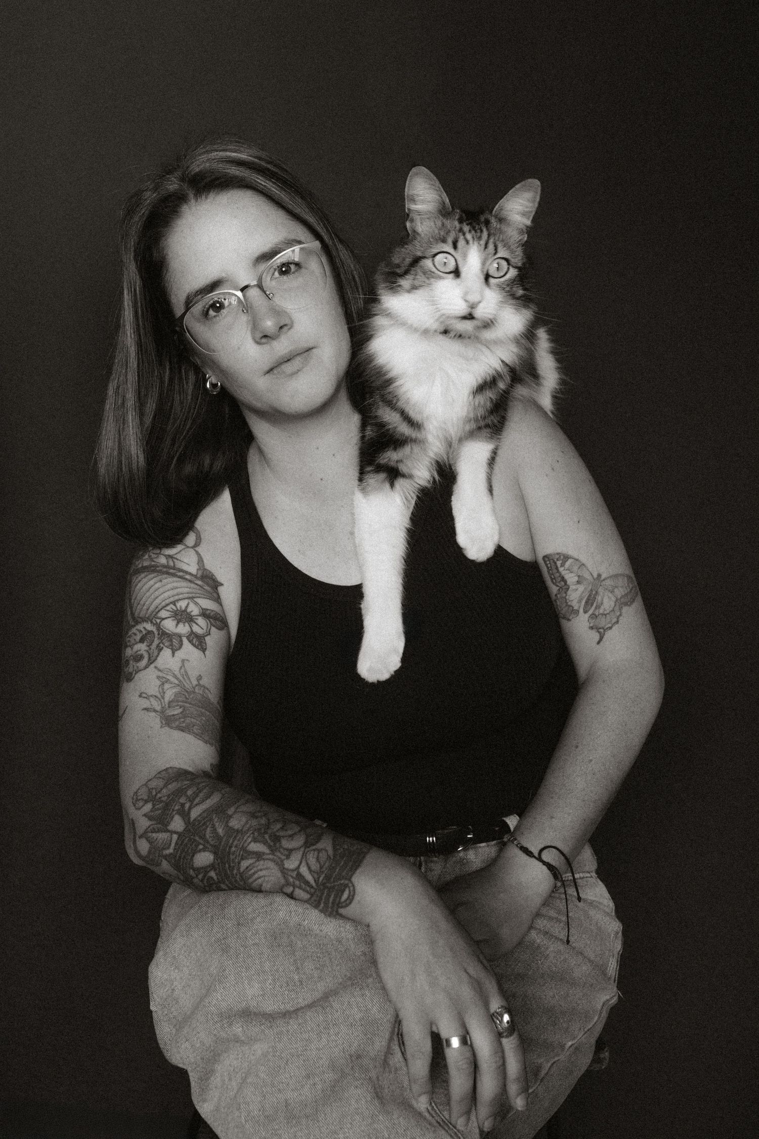 Black and white portrait of a tattooed woman wearing glasses holding a fluffy cat on her shoulder.