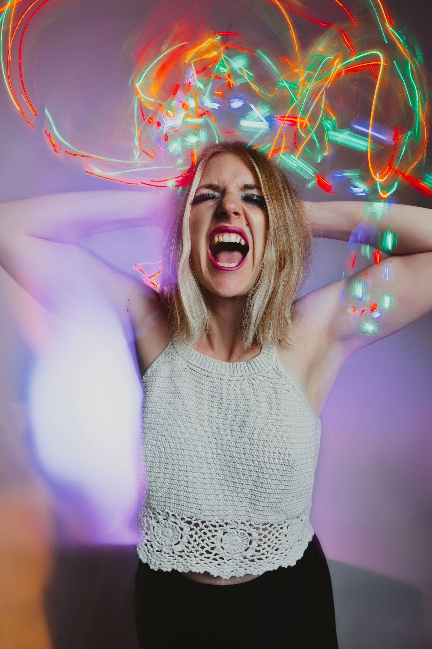 Blonde woman screaming with hands behind head, surrounded by colorful neon light painting trails in a vibrant long exposure photo.