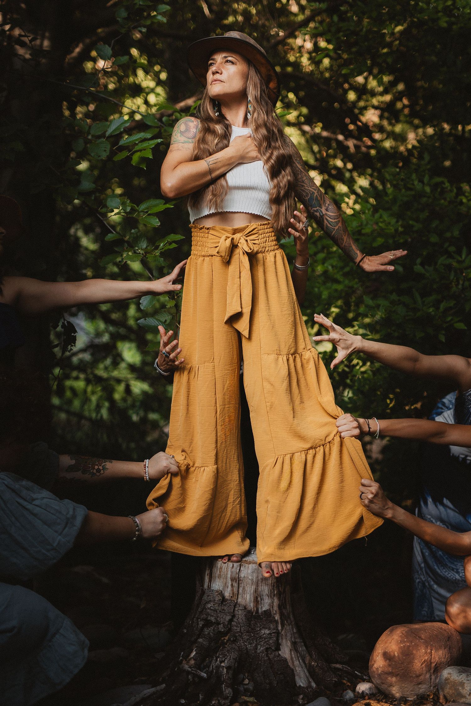 Woman in boho style dress and hat being supported by hidden hands in a whimsical forest photoshoot.