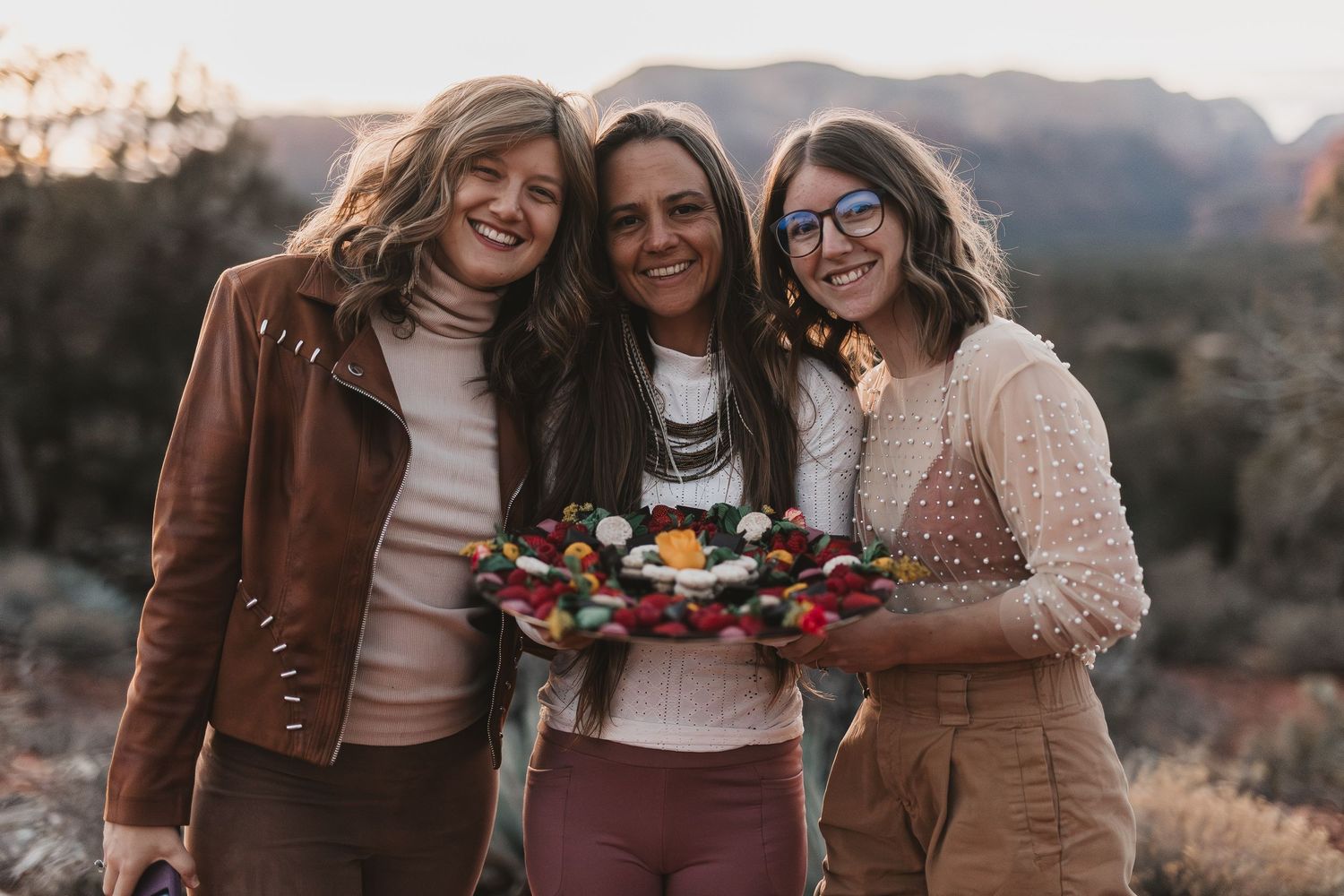 Three friends stand together outdoors at sunset holding a colorful platter of food in a rustic desert setting.