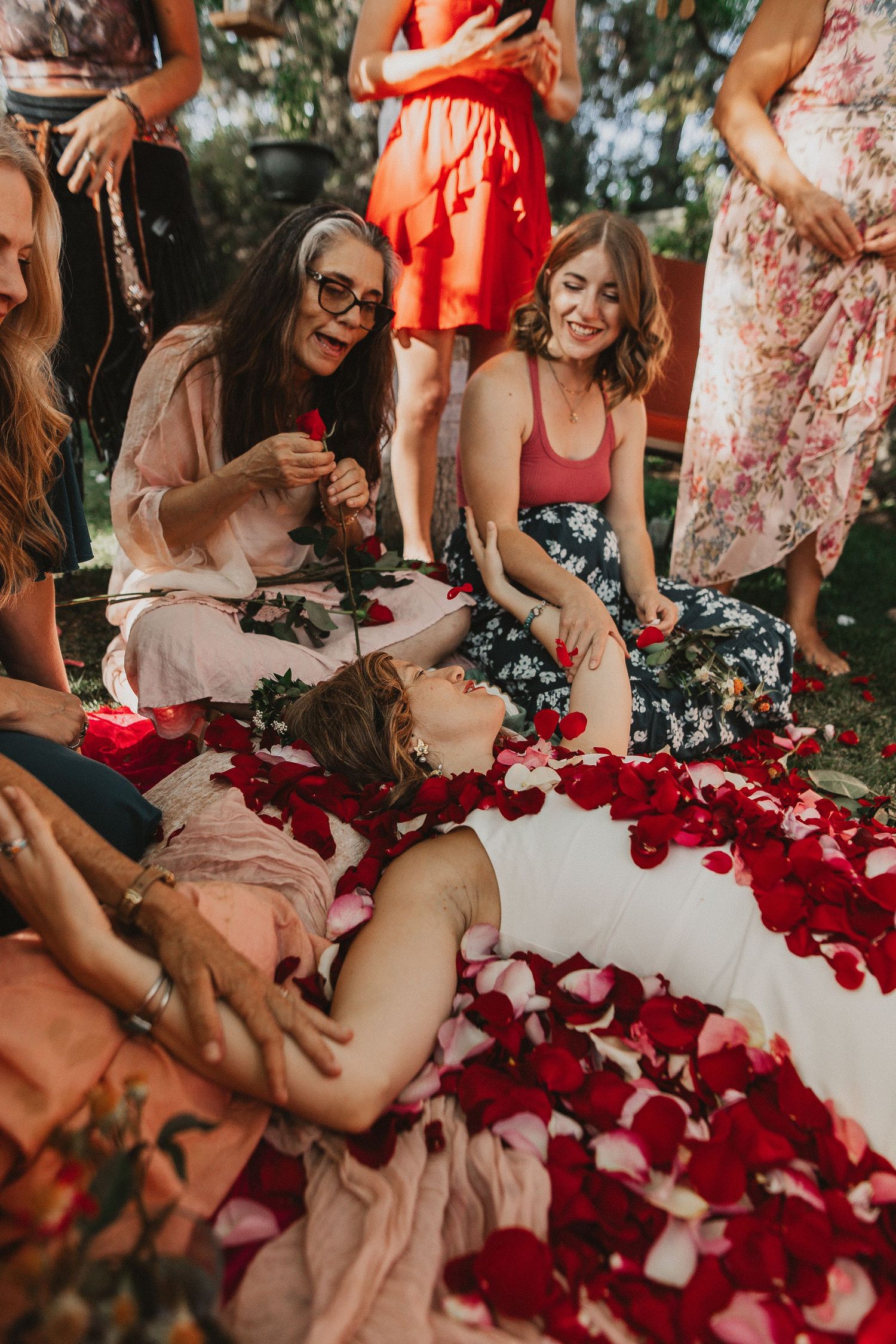 A group of friends laugh and celebrate surrounded by scattered red rose petals on the ground during a sunny outdoor gathering.