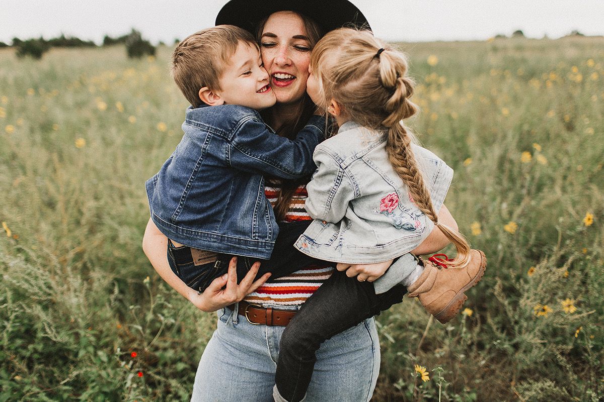 A mother in a black hat holds two young children in a sunny wildflower field, sharing sweet kisses outdoors.