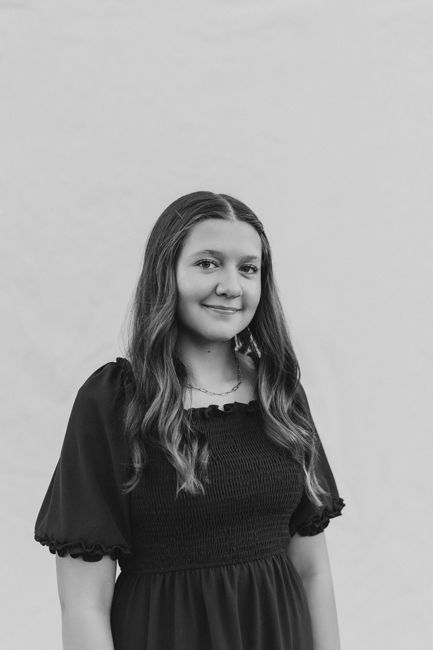Young woman in black smocked dress poses for portrait photo against light background, black and white photography.